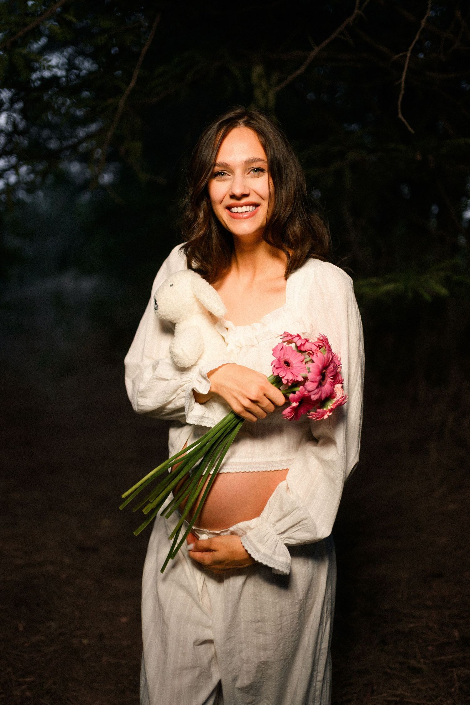 Smiling pregnant woman in white dress holding pink flowers and stuffed bunny outdoors in forest setting.