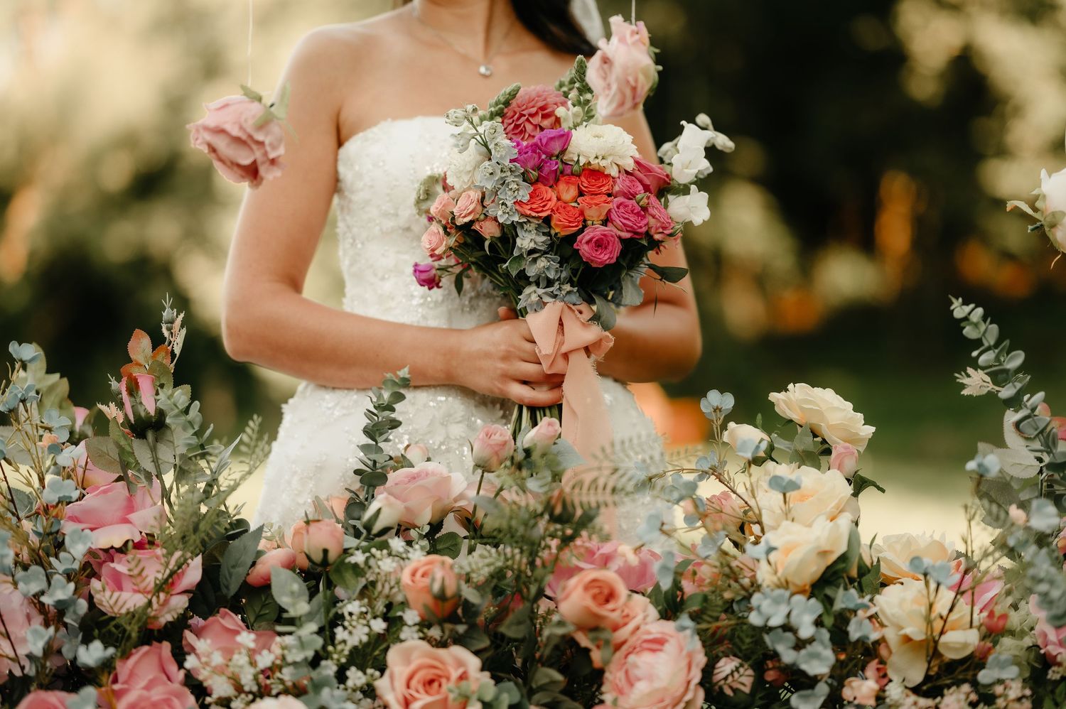 Bride in strapless lace gown holding colorful pink and red bouquet surrounded by romantic floral arrangements.