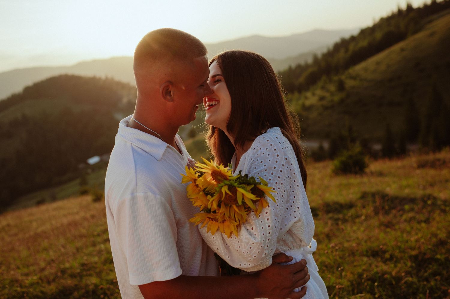 Couple almost kissing at golden hour in mountains, woman holding sunflowers bouquet.
