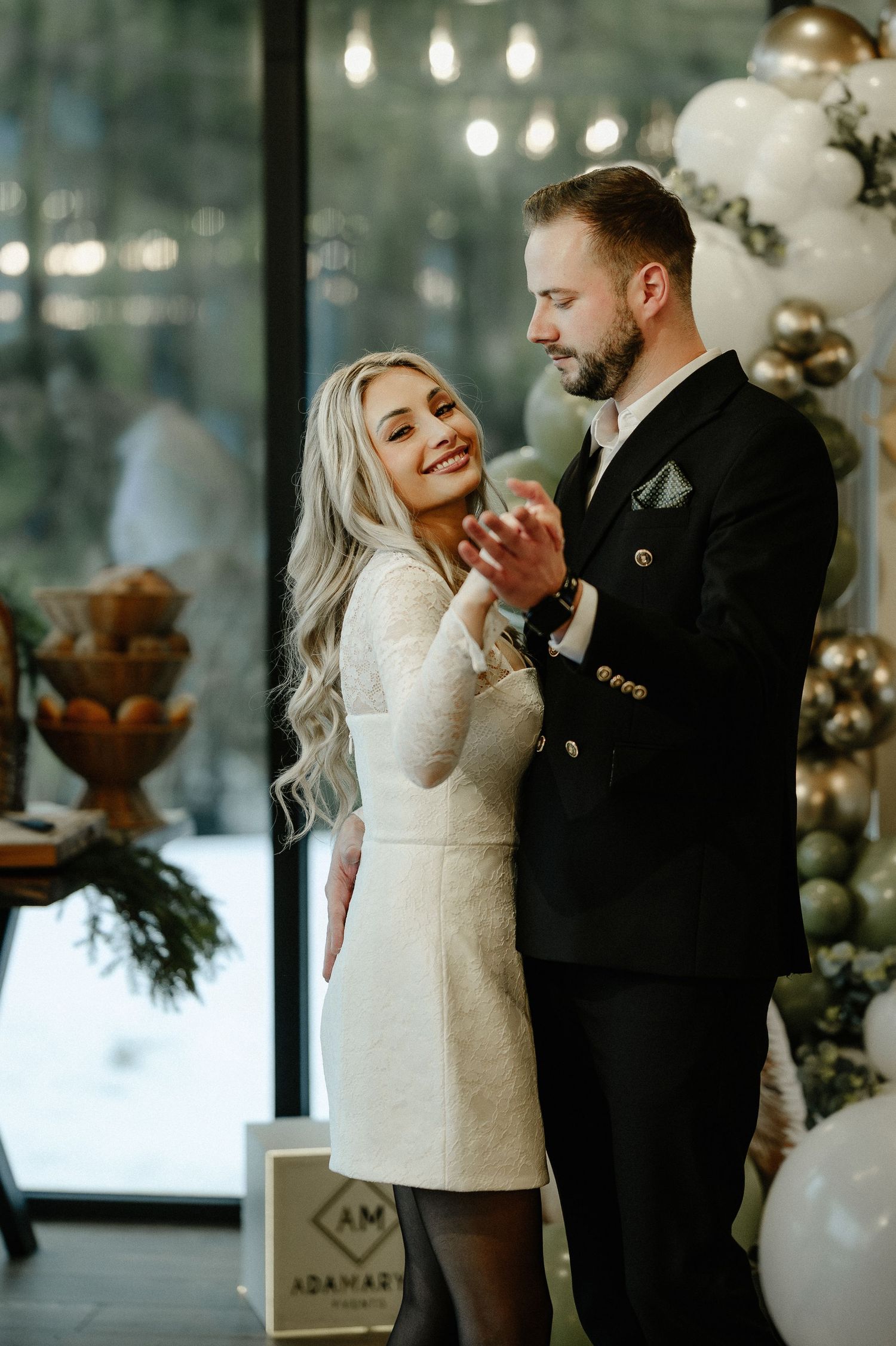 Elegant couple sharing a tender moment at a woodland-themed christening party with white and gold balloon decor.