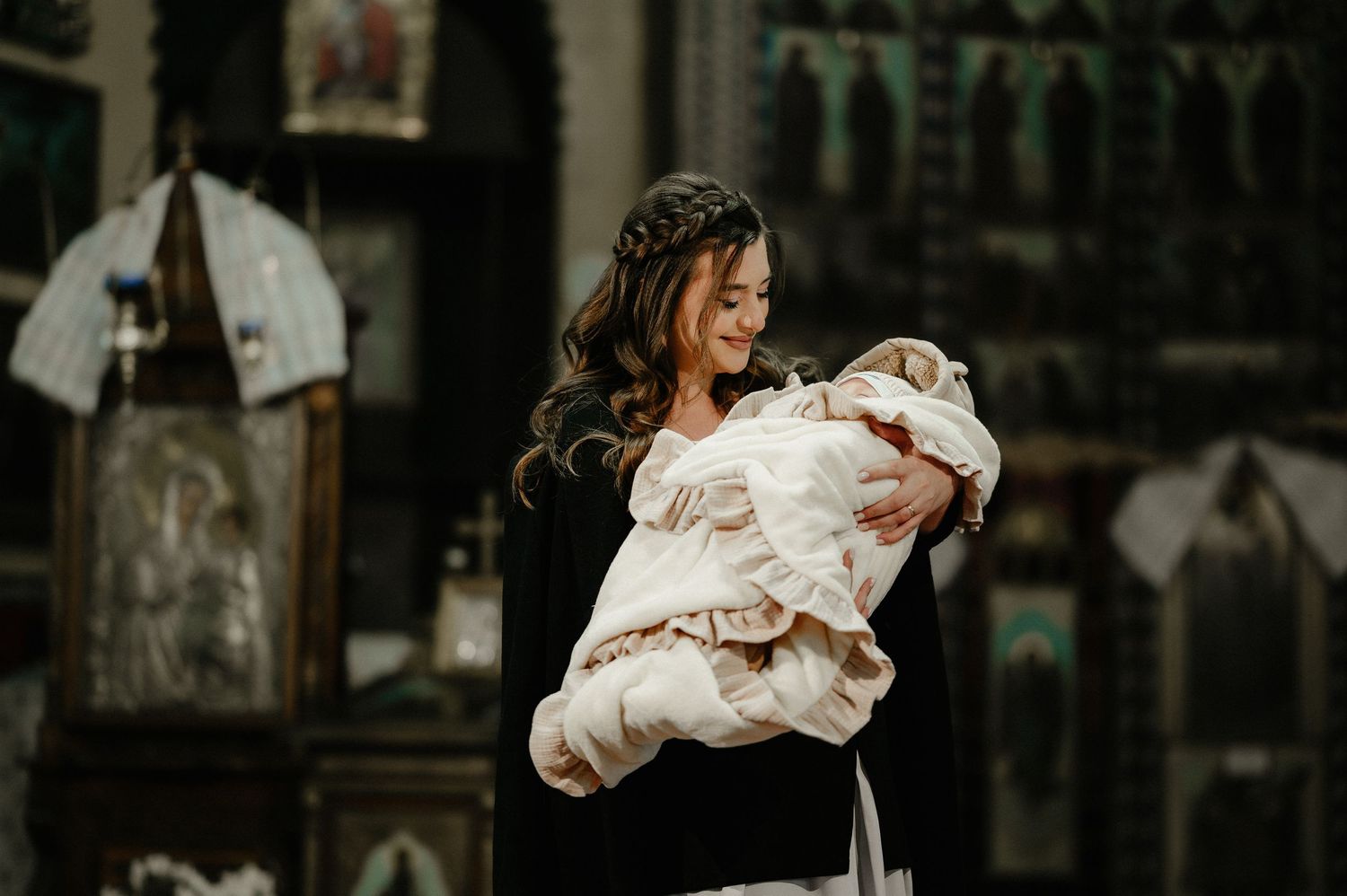 Smiling mother in black dress holding baby in white baptism gown inside Orthodox church.
