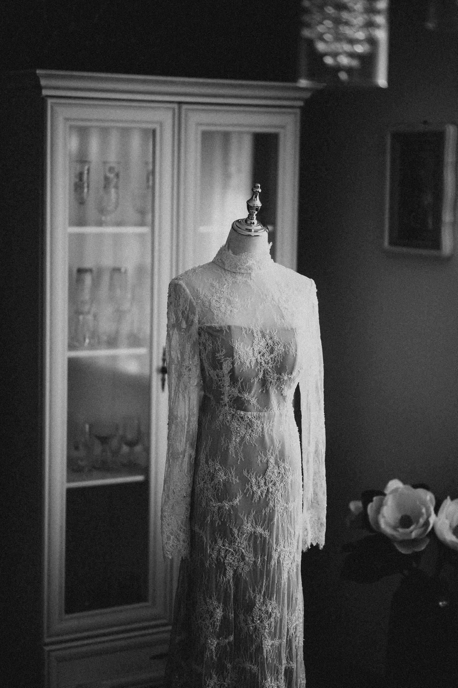 Vintage lace wedding dress displayed on a mannequin in front of an antique white cabinet in black and white.