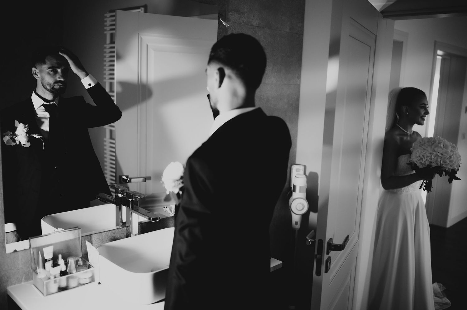 Black and white photo of groom adjusting hair in bathroom mirror as bride stands in doorway.