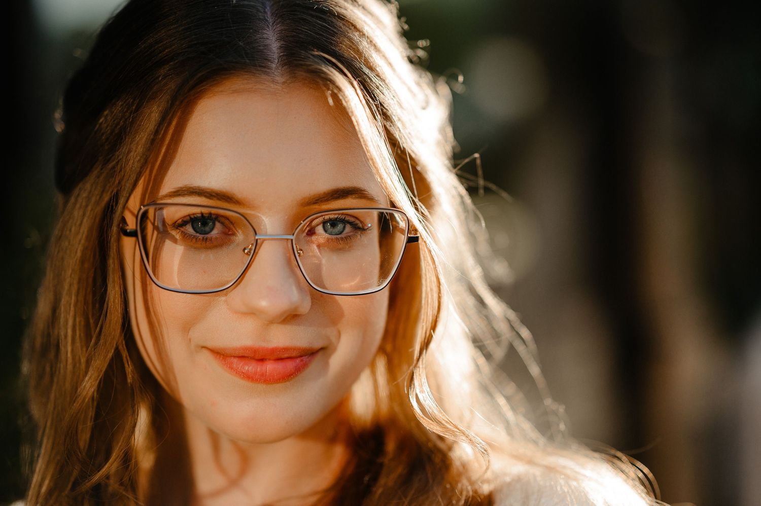 Close-up portrait of young woman with glasses and wavy hair in warm golden sunlight.