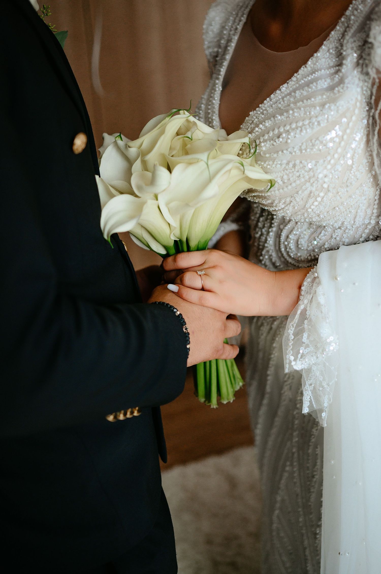 Close-up of bride and groom holding white calla lily bouquet, bride wearing beaded wedding dress with gold ring.