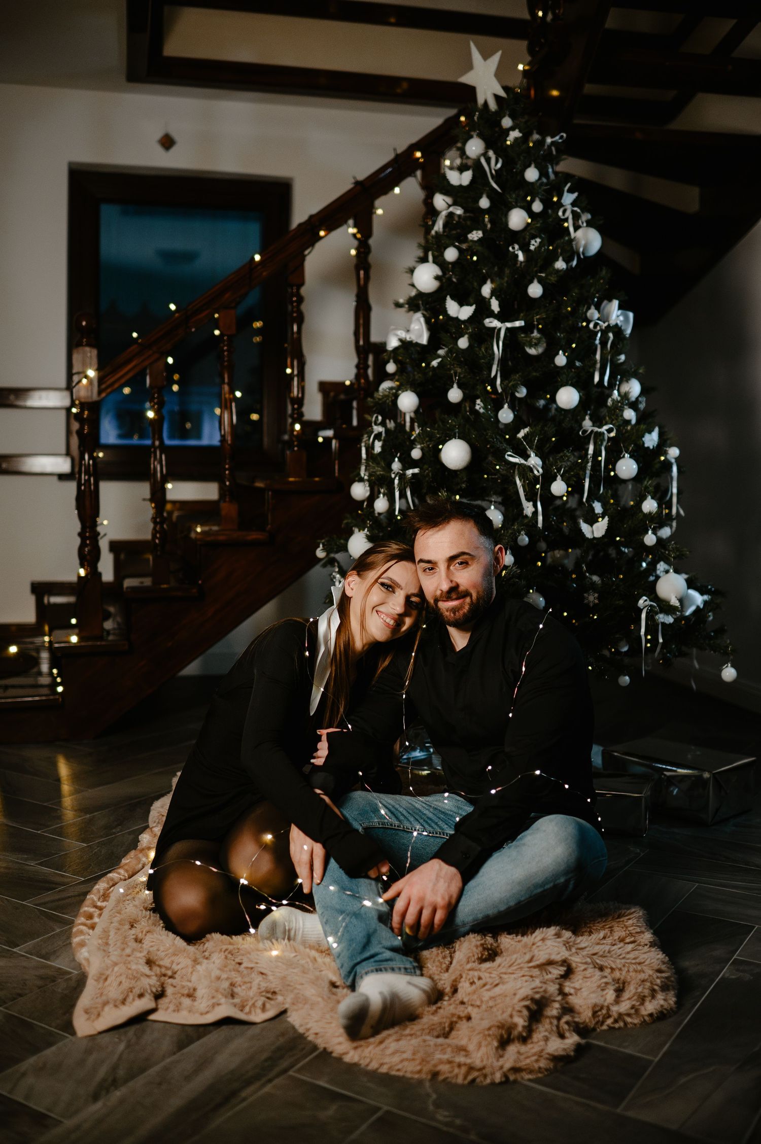 Couple sitting on a fur rug in front of a lit Christmas tree holding gold ornaments.