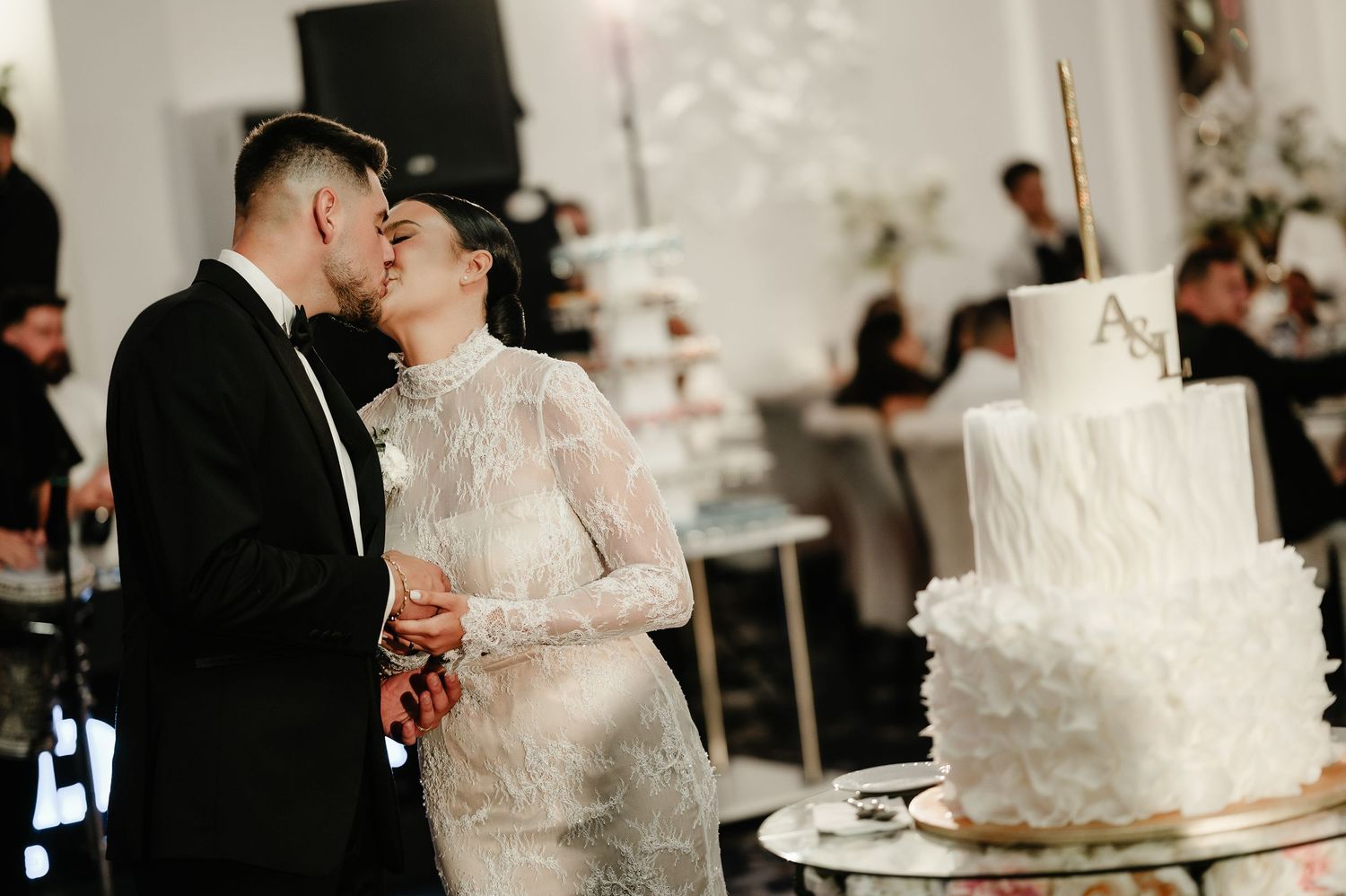 Bride and groom sharing a kiss beside a stunning white two-tier ruffled wedding cake.