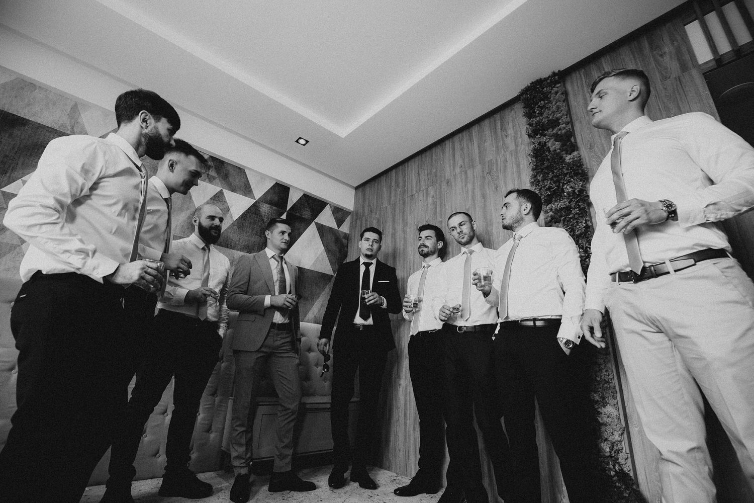 Black and white photo of groom and groomsmen gathered in circle holding drinks during wedding preparations.