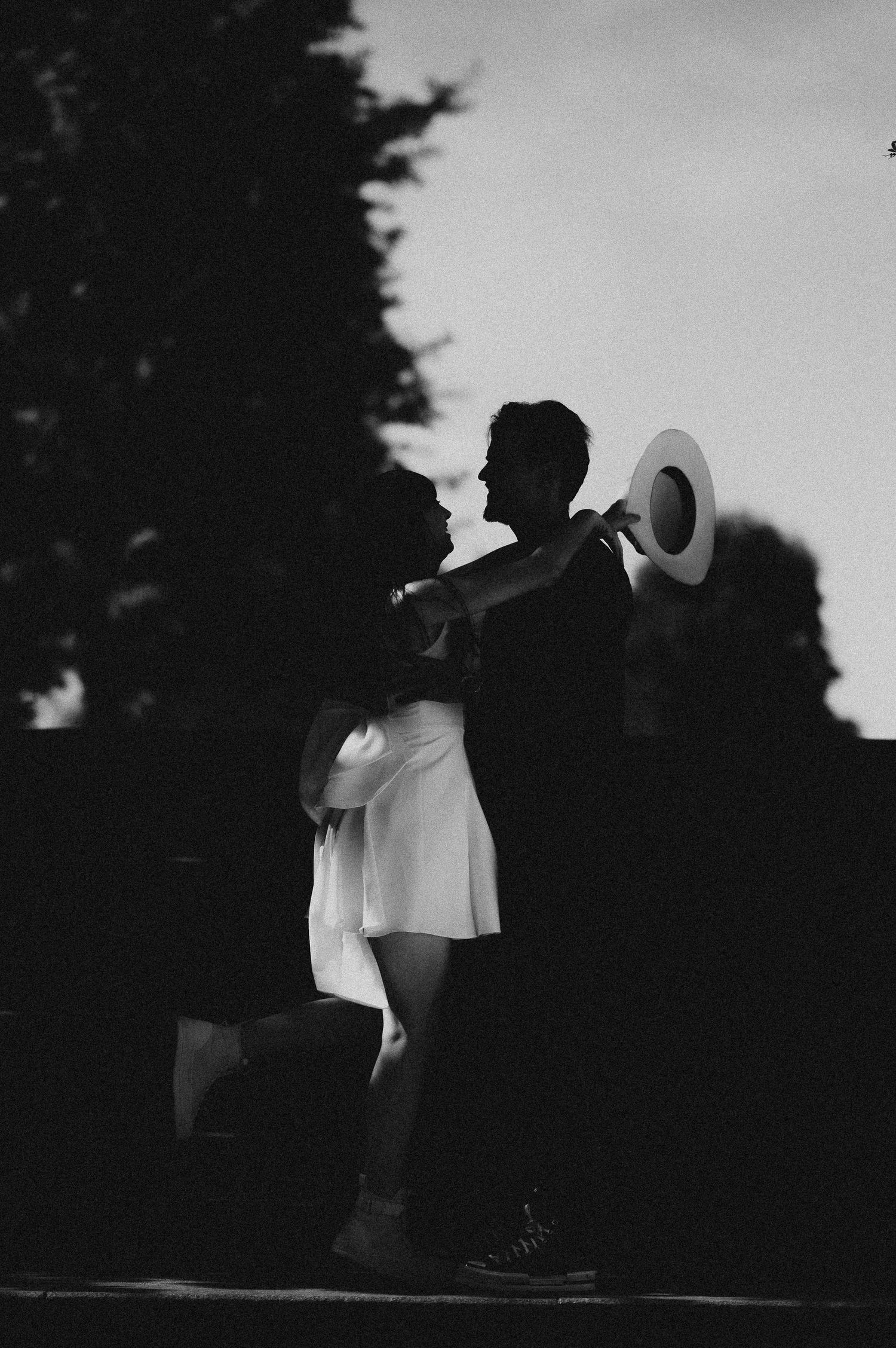 Black and white silhouette of a romantic couple embracing at dusk, woman holding a wide-brim hat.