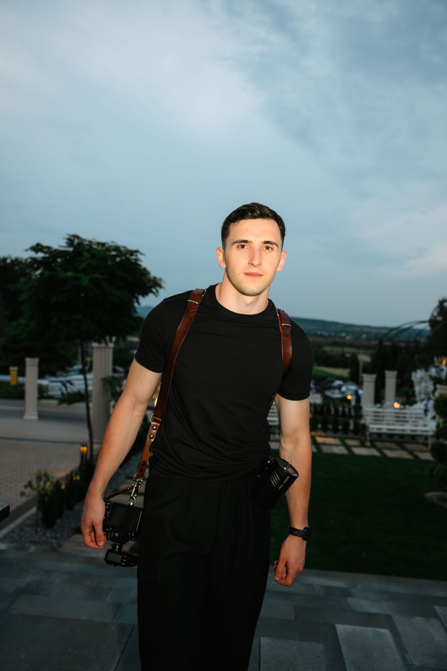 Young man in black t-shirt and pants carrying a camera on a strap, standing outdoors against a cloudy sky.