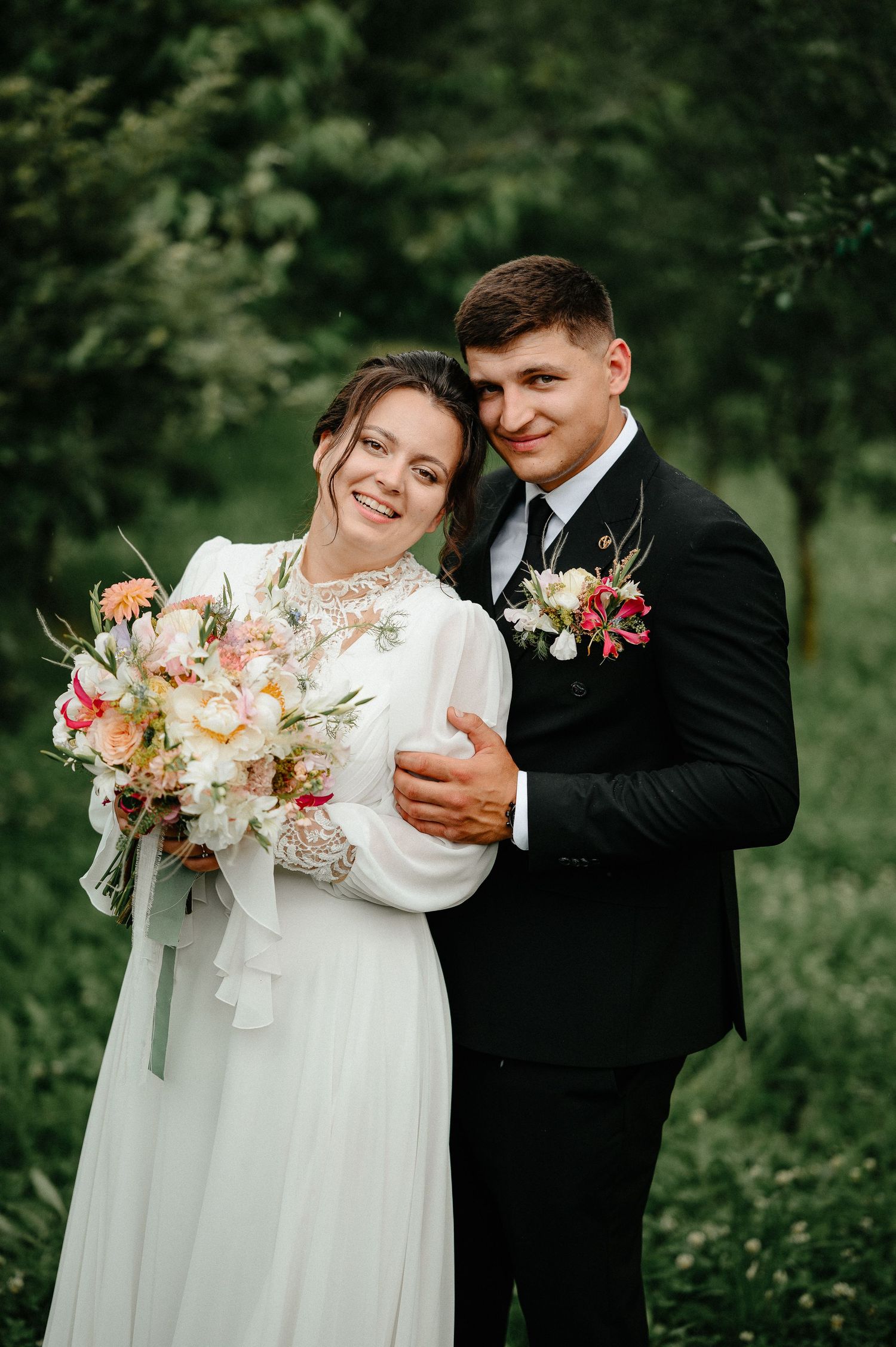 A smiling bride in a white dress holding a bouquet stands with a groom in a black suit outdoors.