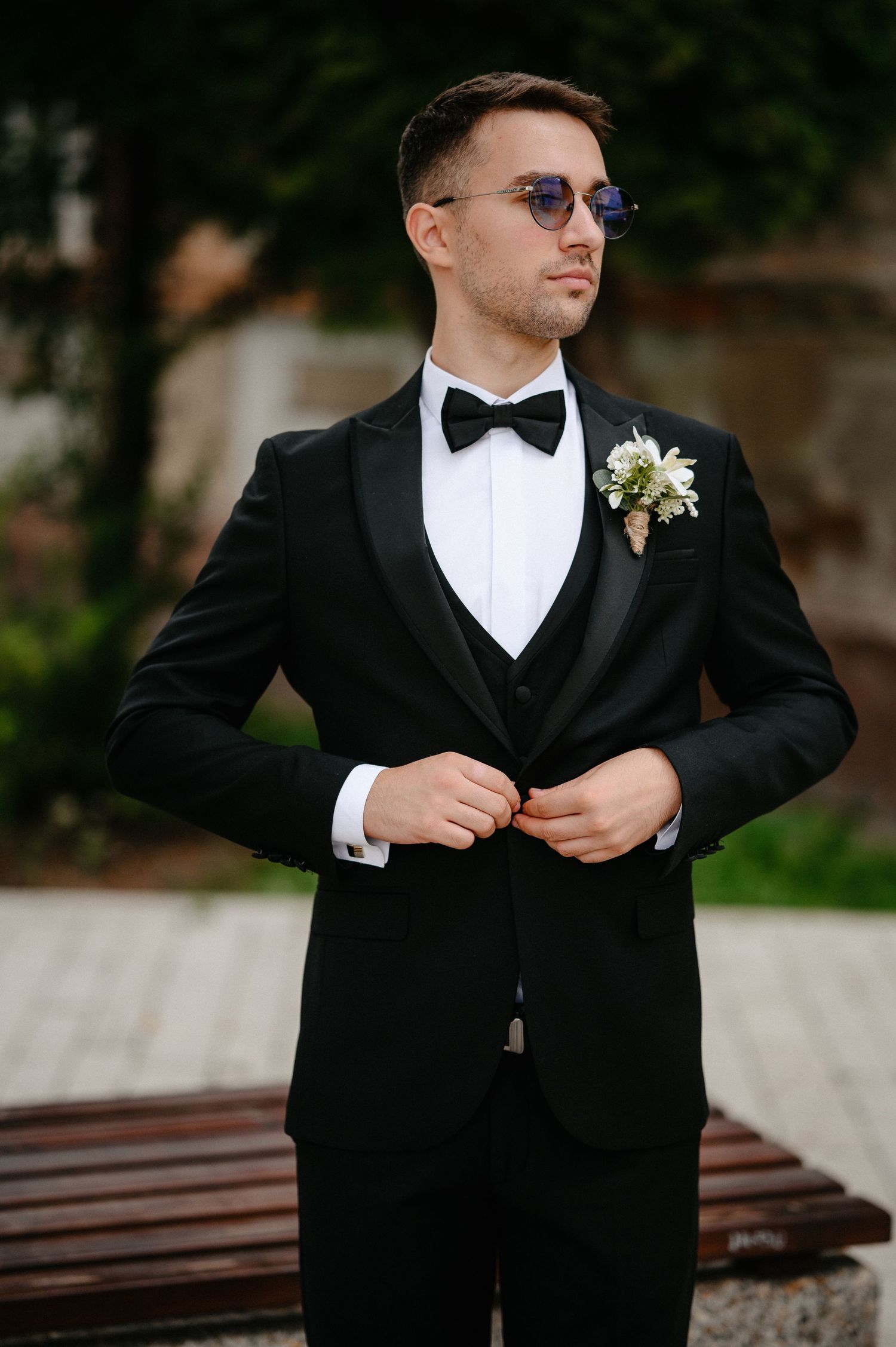Elegant groom in black tuxedo with bow tie and boutonniere posing outdoors on wedding day.