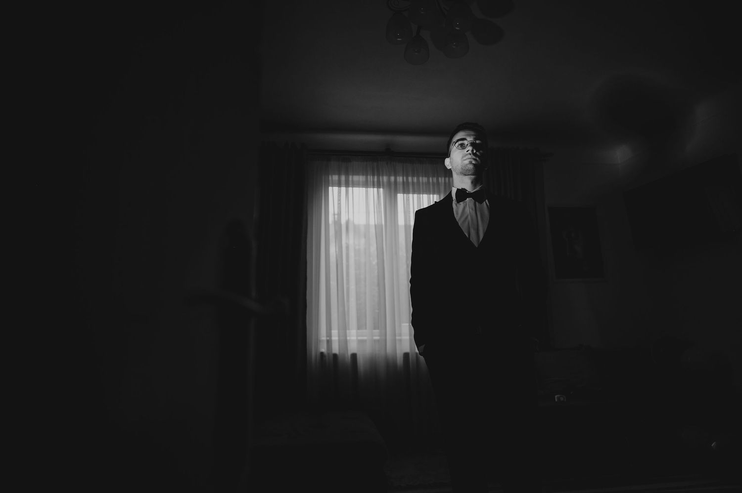 A groom in a black suit and bow tie stands silhouetted by a curtained window in a moody black and white portrait.