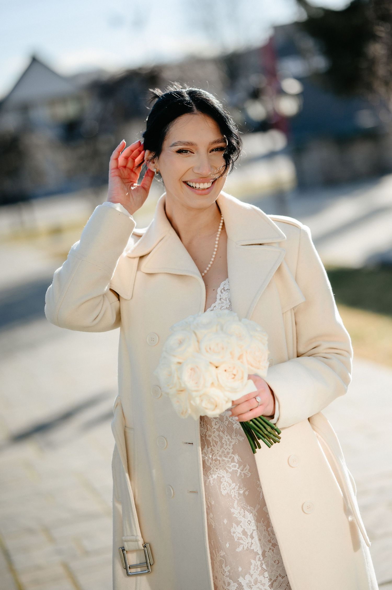 Smiling bride in beige trench coat holding white rose bouquet outdoors on sunny day.