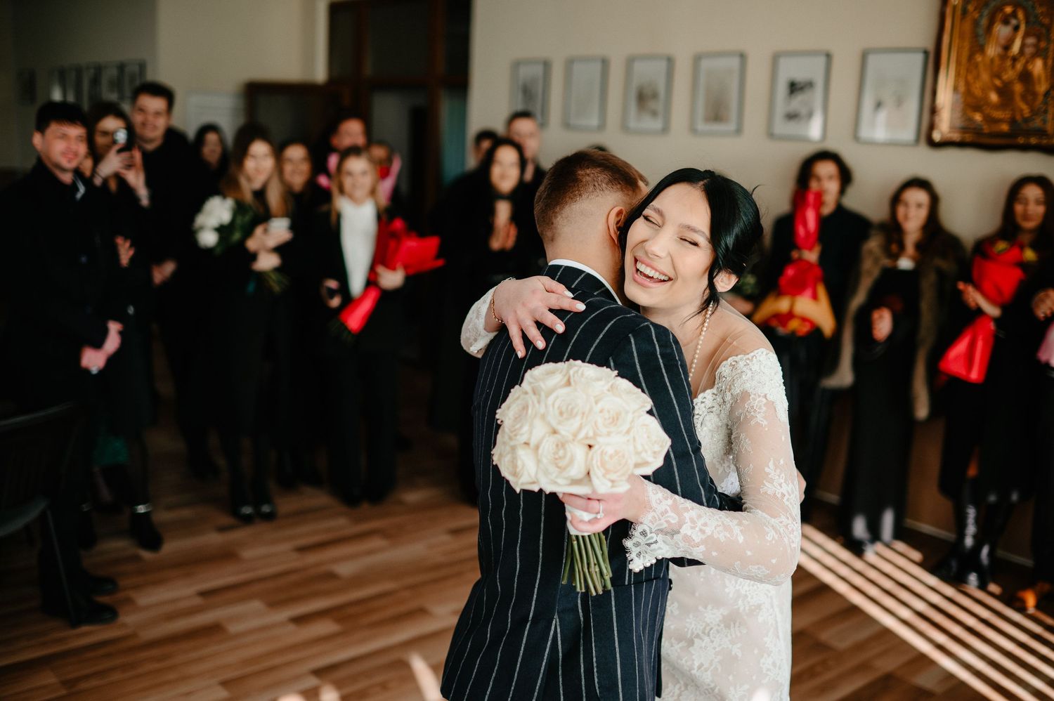 Bride smiling and hugging groom at wedding ceremony, holding white bouquet, guests in background holding red flowers.