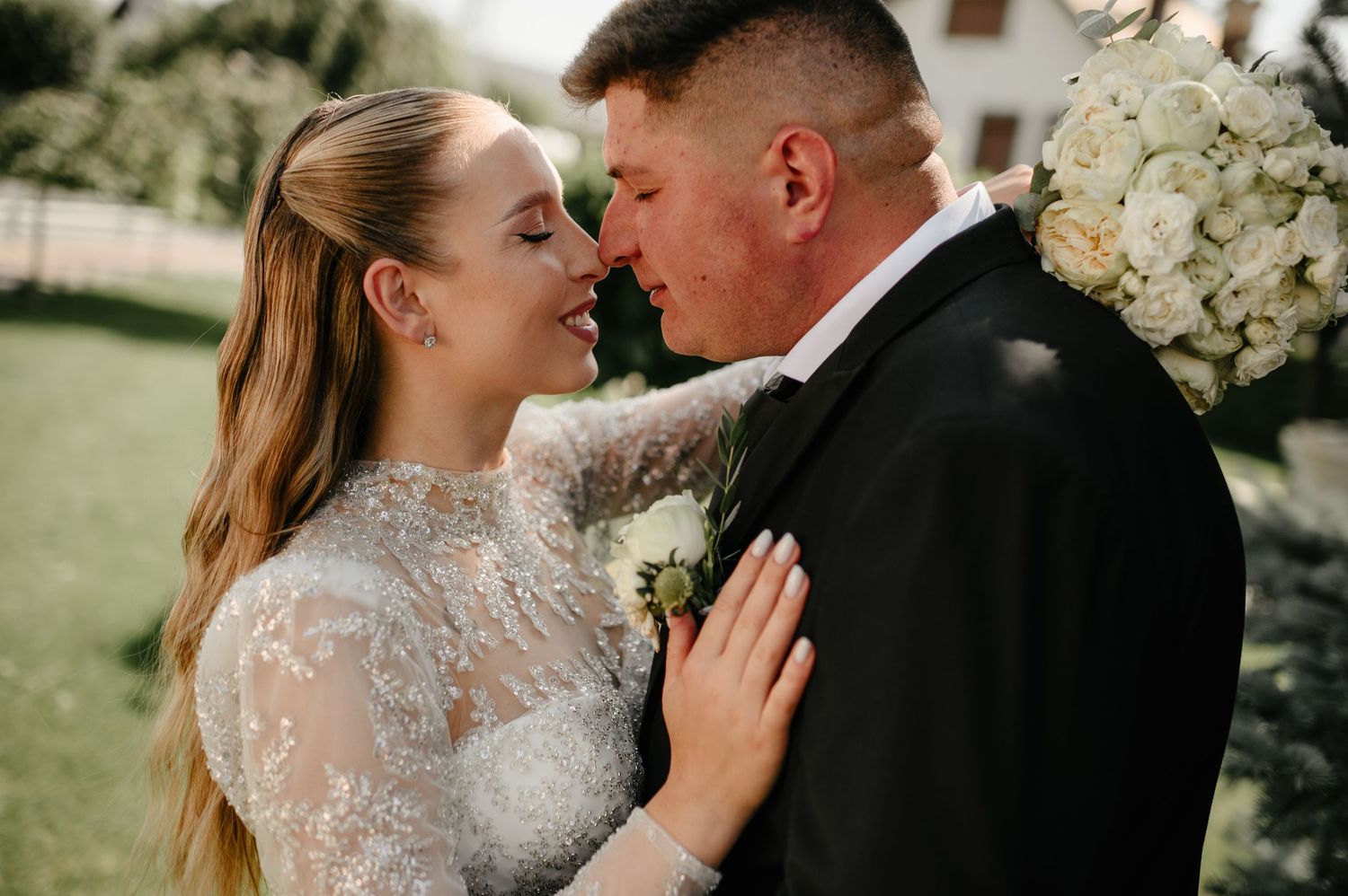 Smiling bride in beaded lace dress embracing groom in black tuxedo near white floral arrangement.