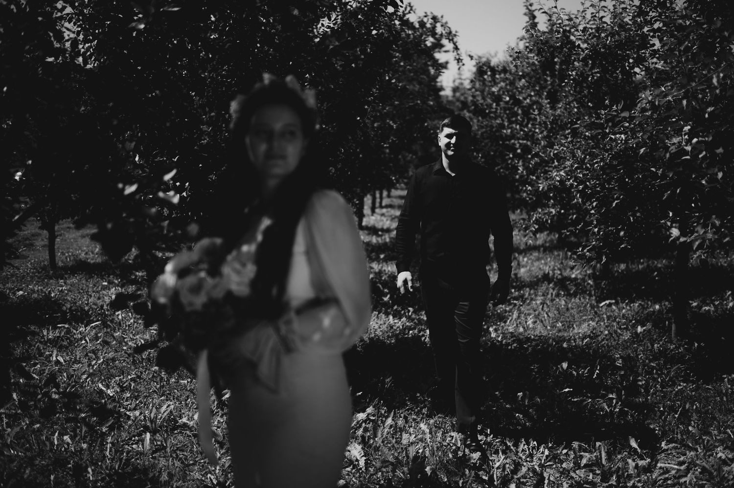 Black and white photo of bride holding bouquet with groom walking through orchard trees in background.