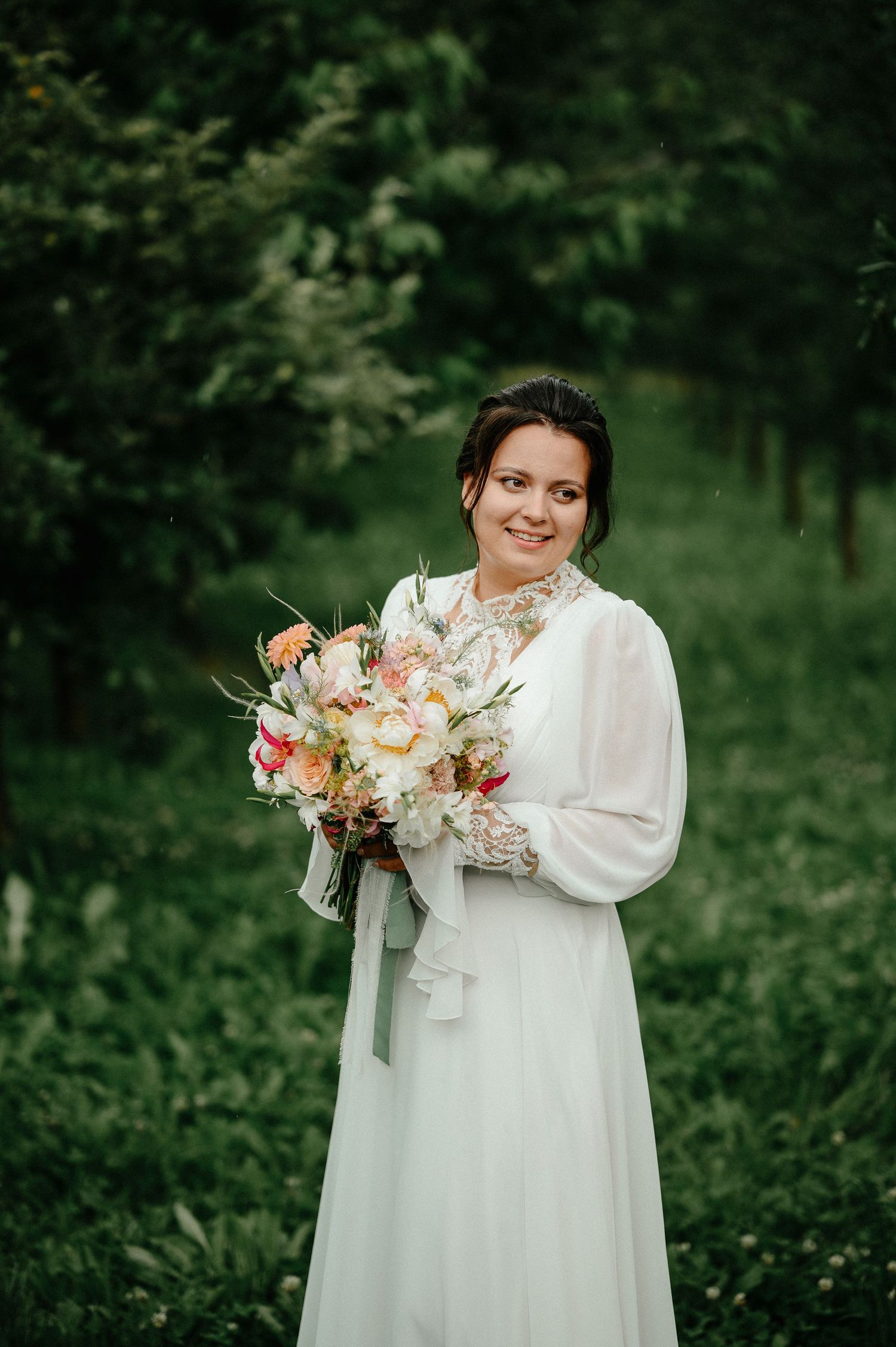 Bride in white long-sleeve gown holding colorful bouquet smiling in lush green garden setting.