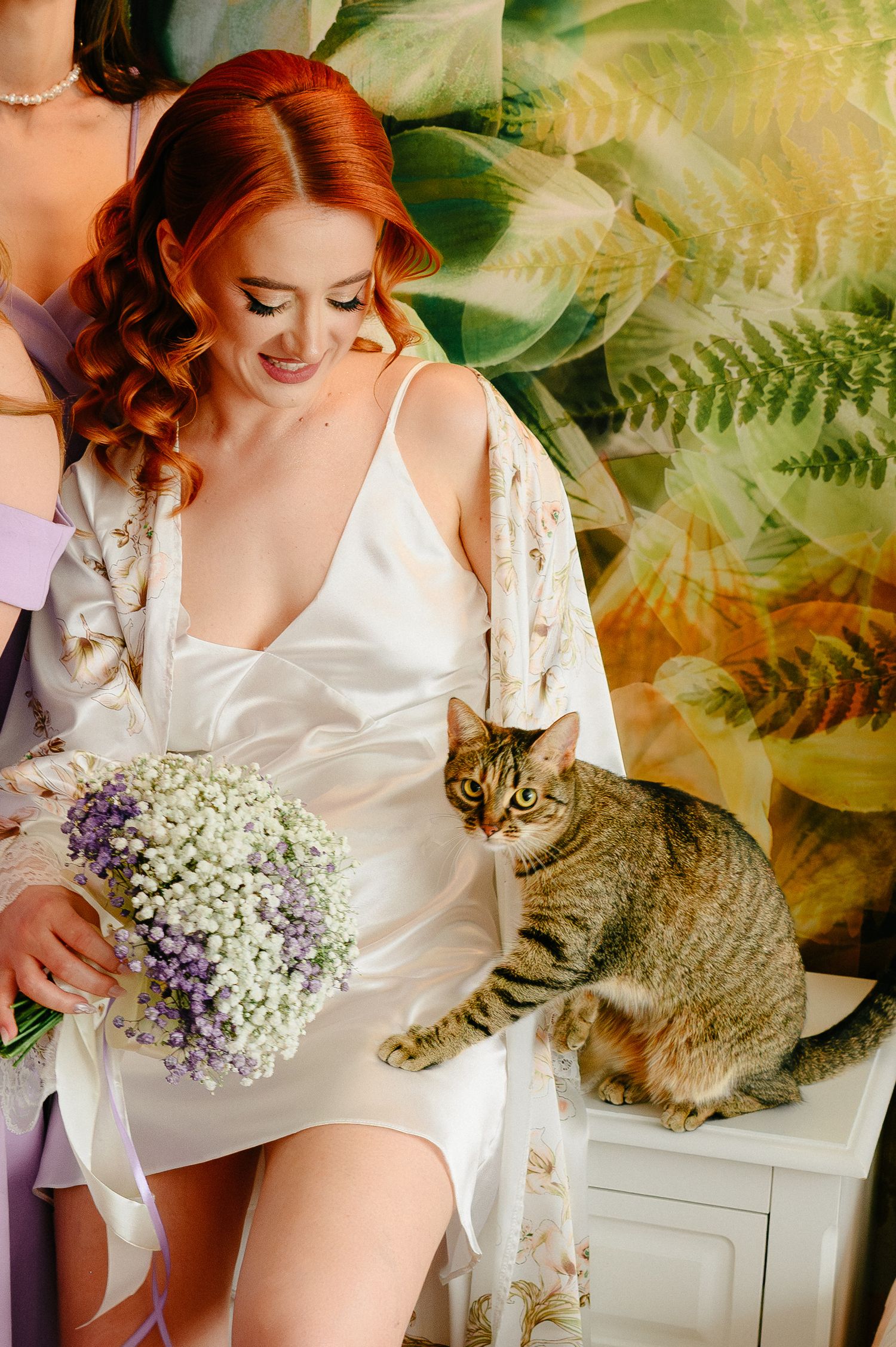 Red-haired bride in white satin robe holds purple floral bouquet while tabby cat sits beside her against tropical leaf backdrop.