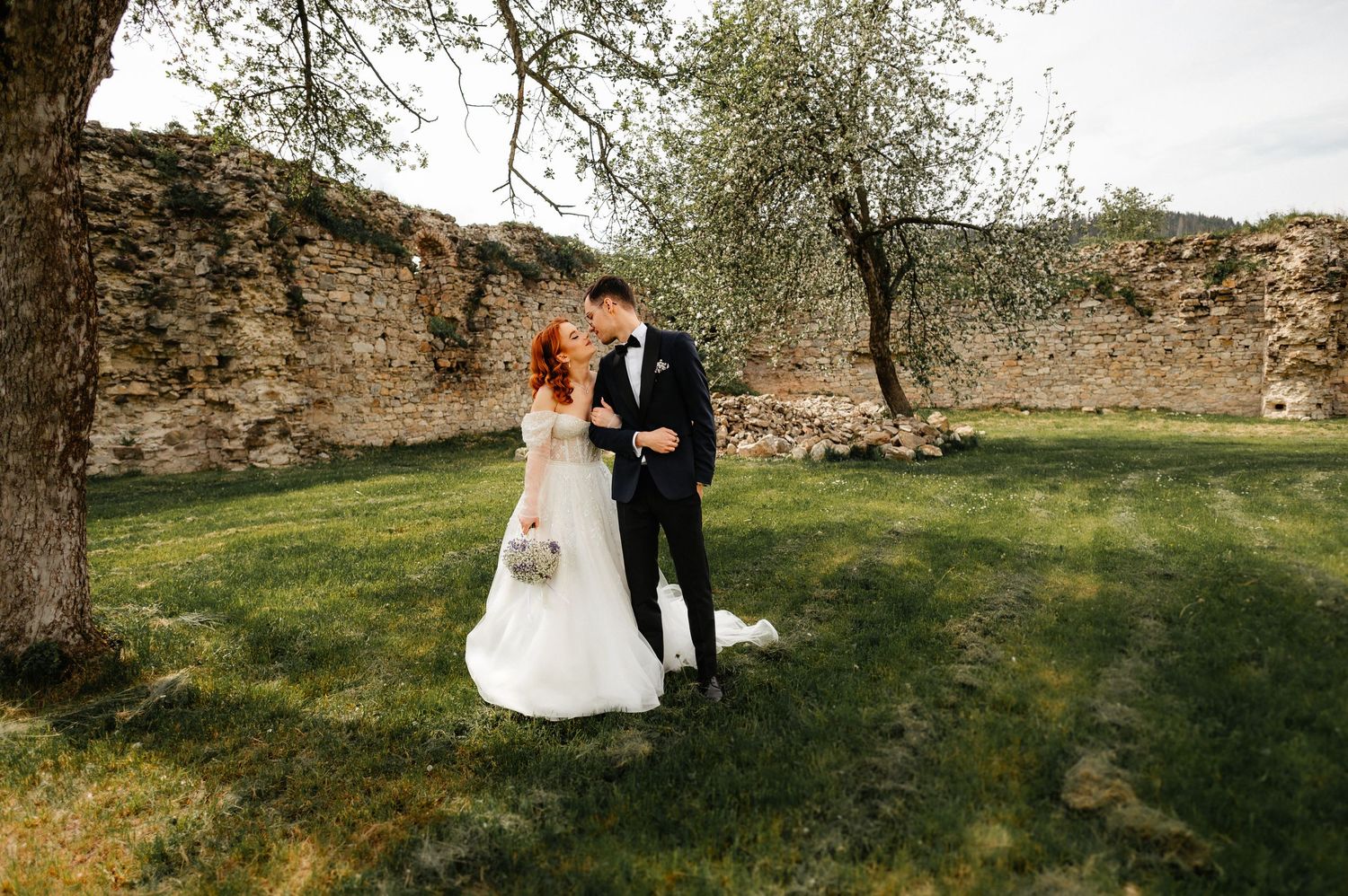A bride and groom share a romantic kiss on green grass near a rustic stone wall and tree on their wedding day.
