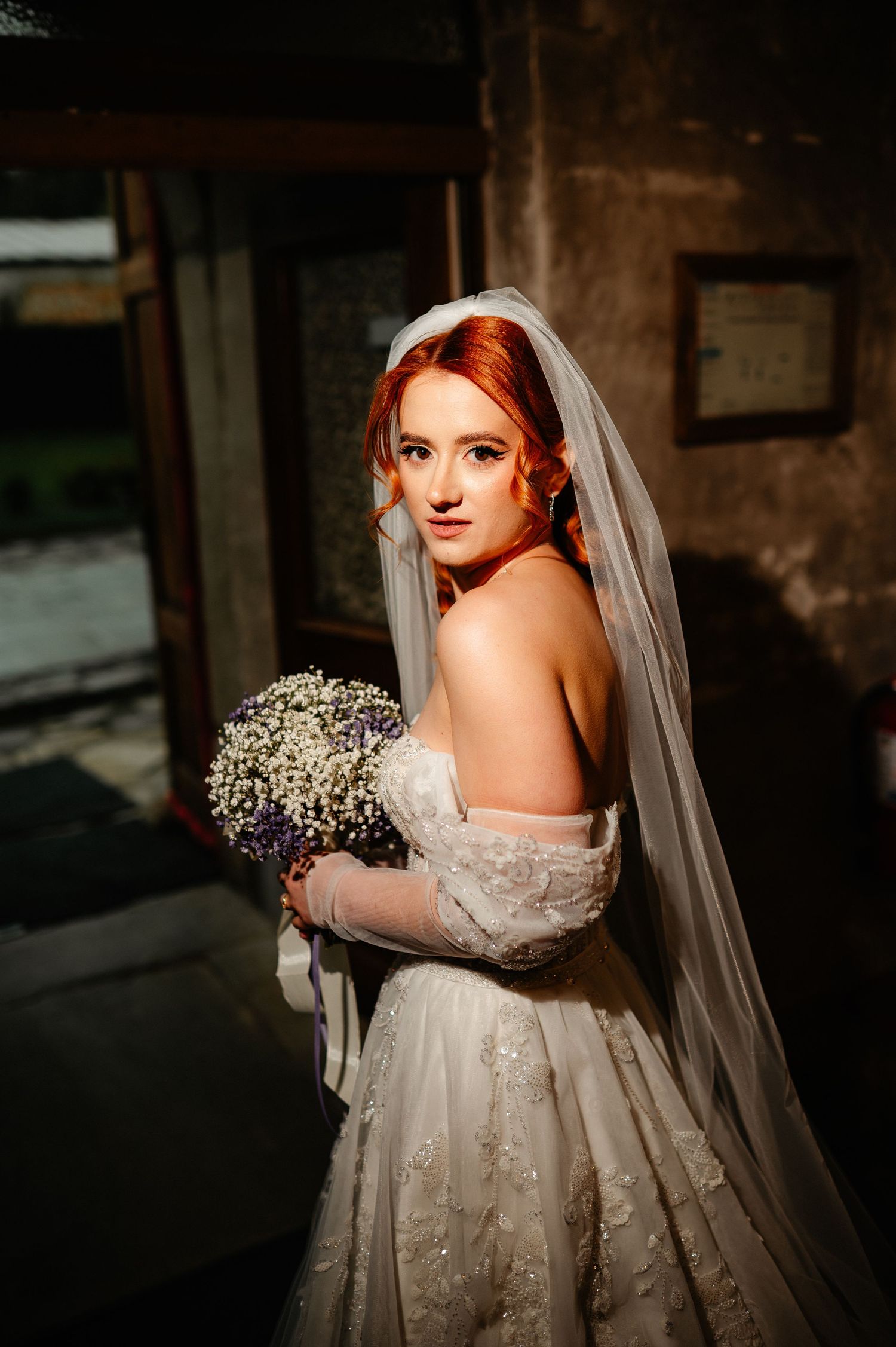 Red-haired bride in off-shoulder gown and veil holds baby's breath bouquet, posing elegantly in warm light.