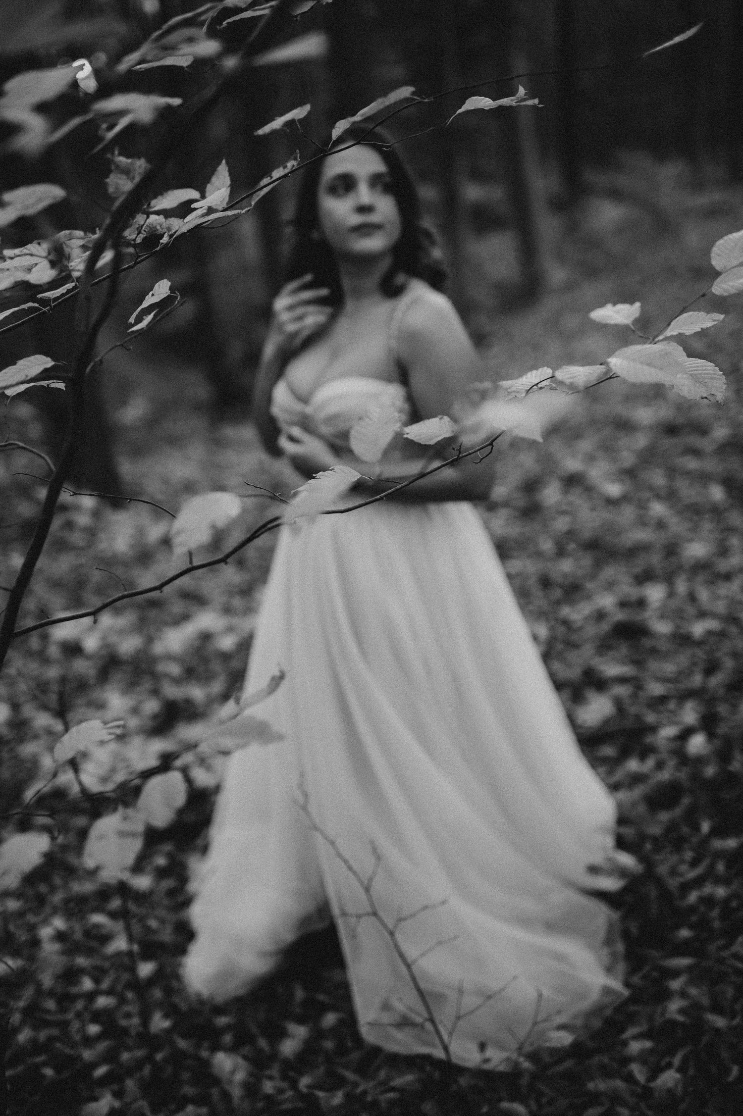 Black and white photo of a woman in a flowing white gown standing in a misty forest surrounded by autumn leaves.