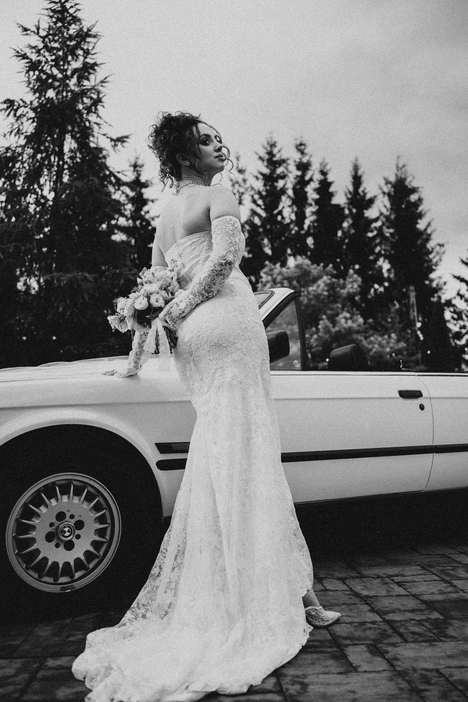 Black and white photo of a bride in a lace gown holding a bouquet beside a white convertible car.