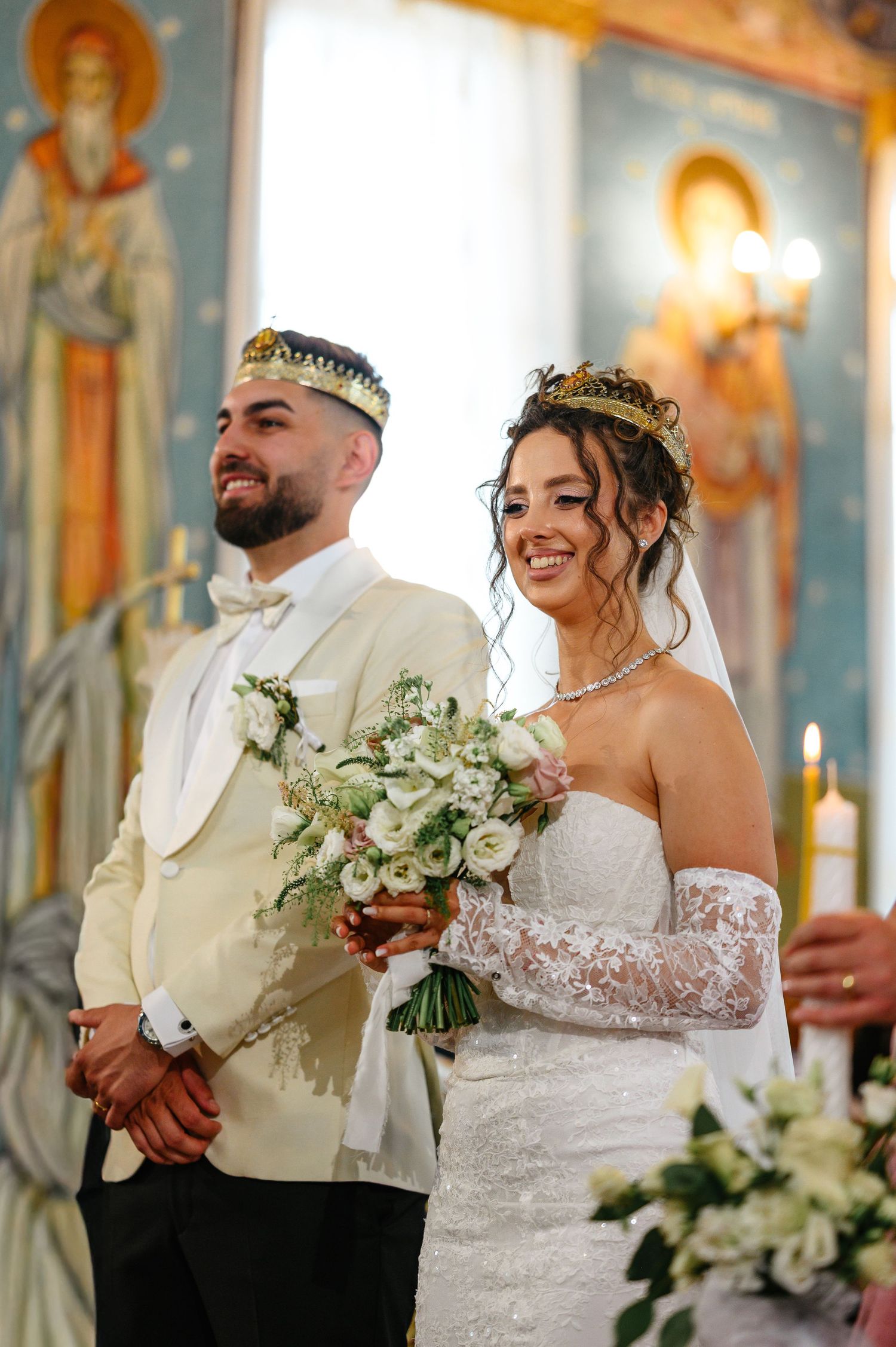 Bride and groom in gold crowns during Orthodox wedding ceremony, bride holding white bouquet in elegant lace gown.