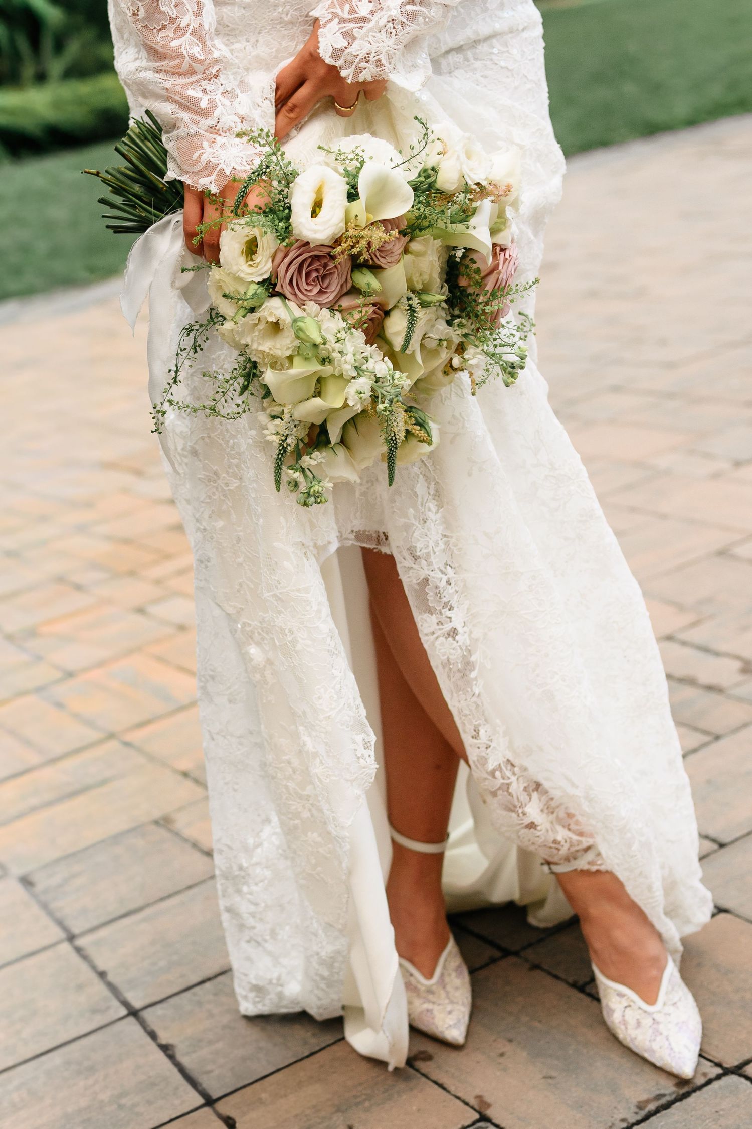 Bride in lace wedding dress holding a bouquet of roses and calla lilies, wearing white lace heels on a stone patio.