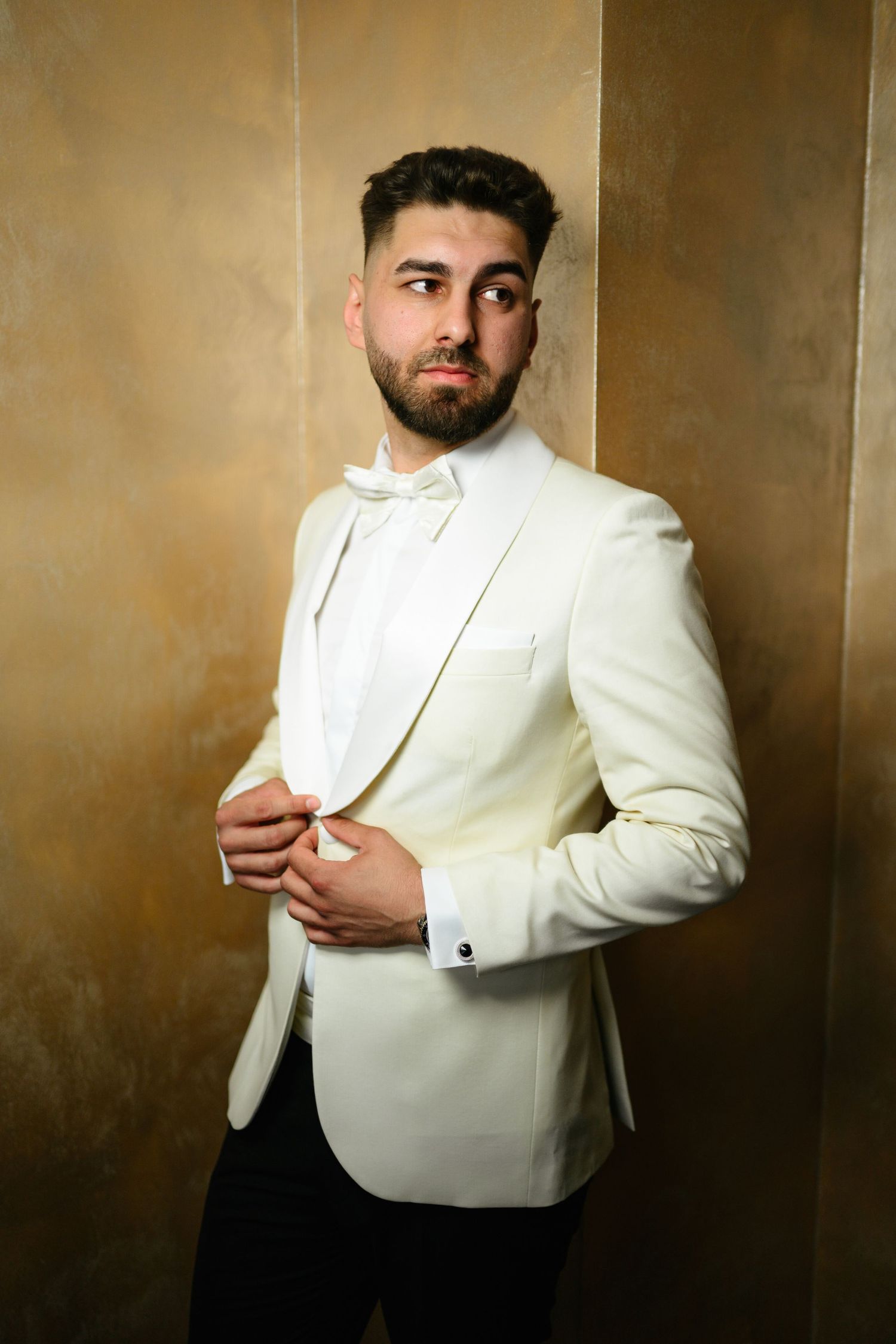 A stylish man in a white tuxedo jacket and bow tie adjusts his button against a golden wall.