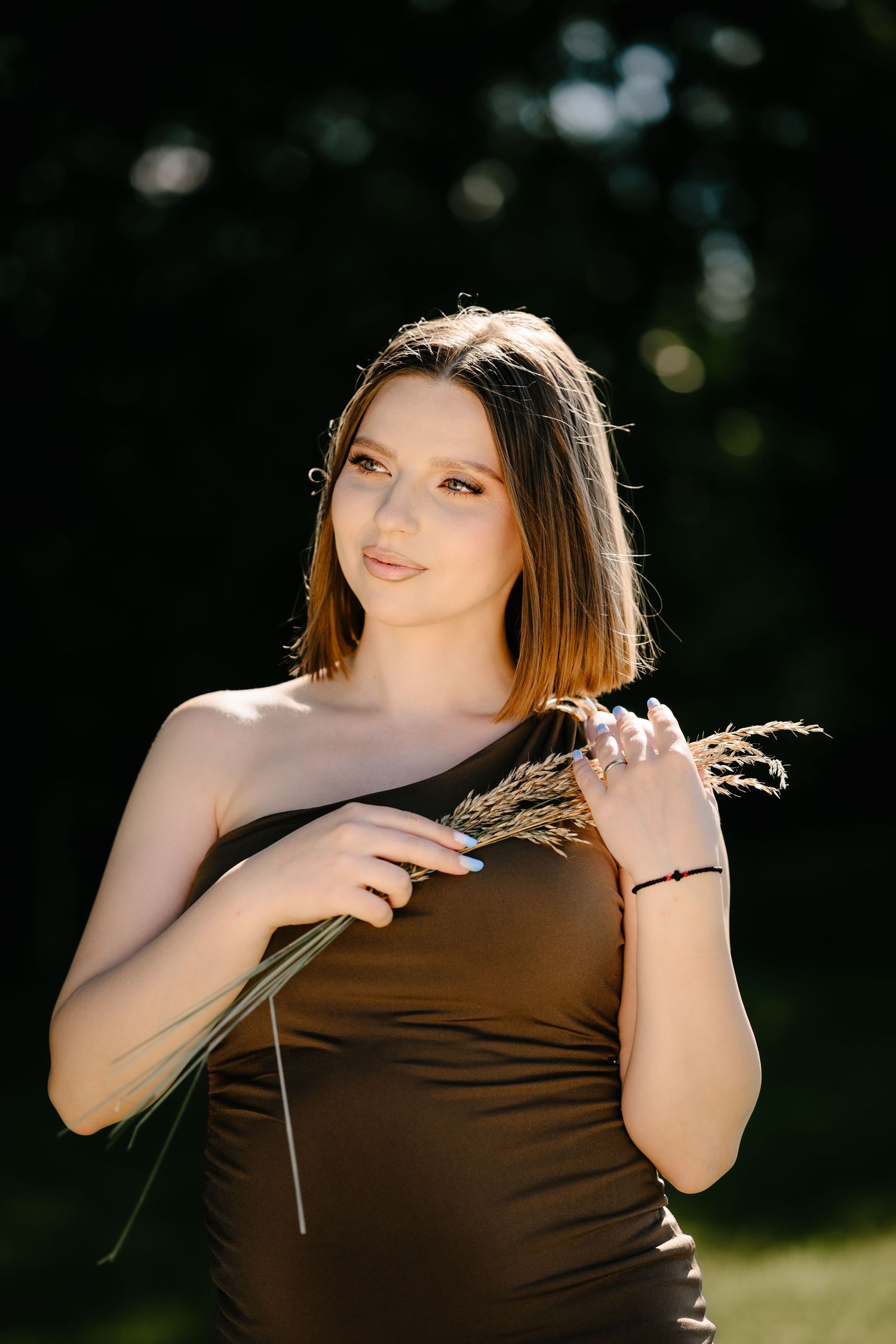 Young woman in brown one-shoulder dress holding dried grass stems in sunlit outdoor portrait.