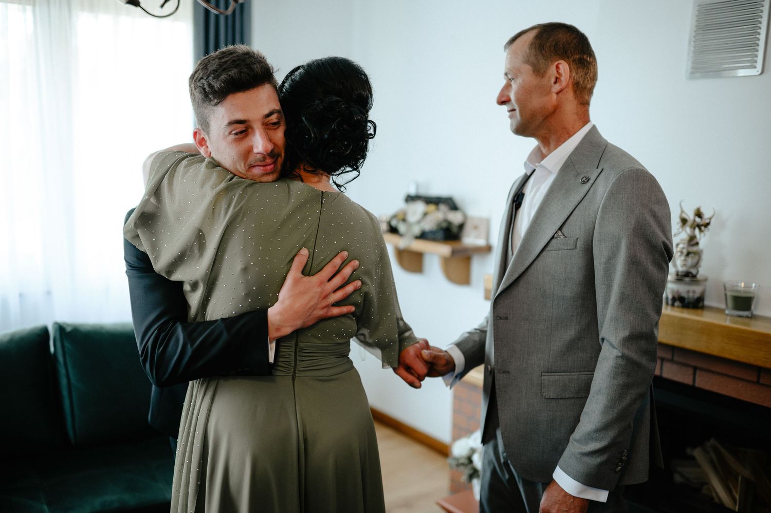 Groom embracing mother while father watches during pre-wedding preparations.