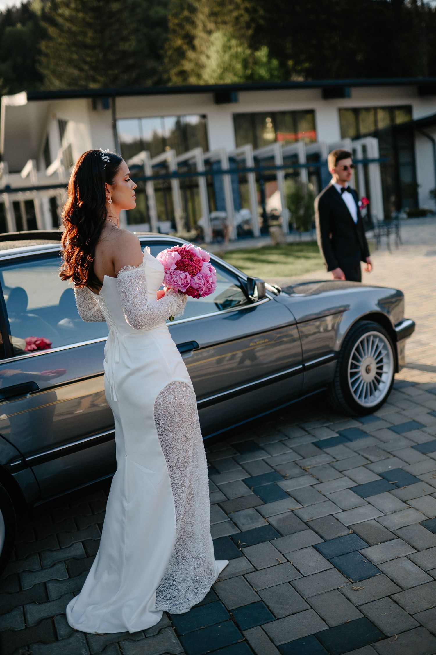 Bride in off-shoulder lace gown holding pink bouquet leans on classic BMW as groom approaches.