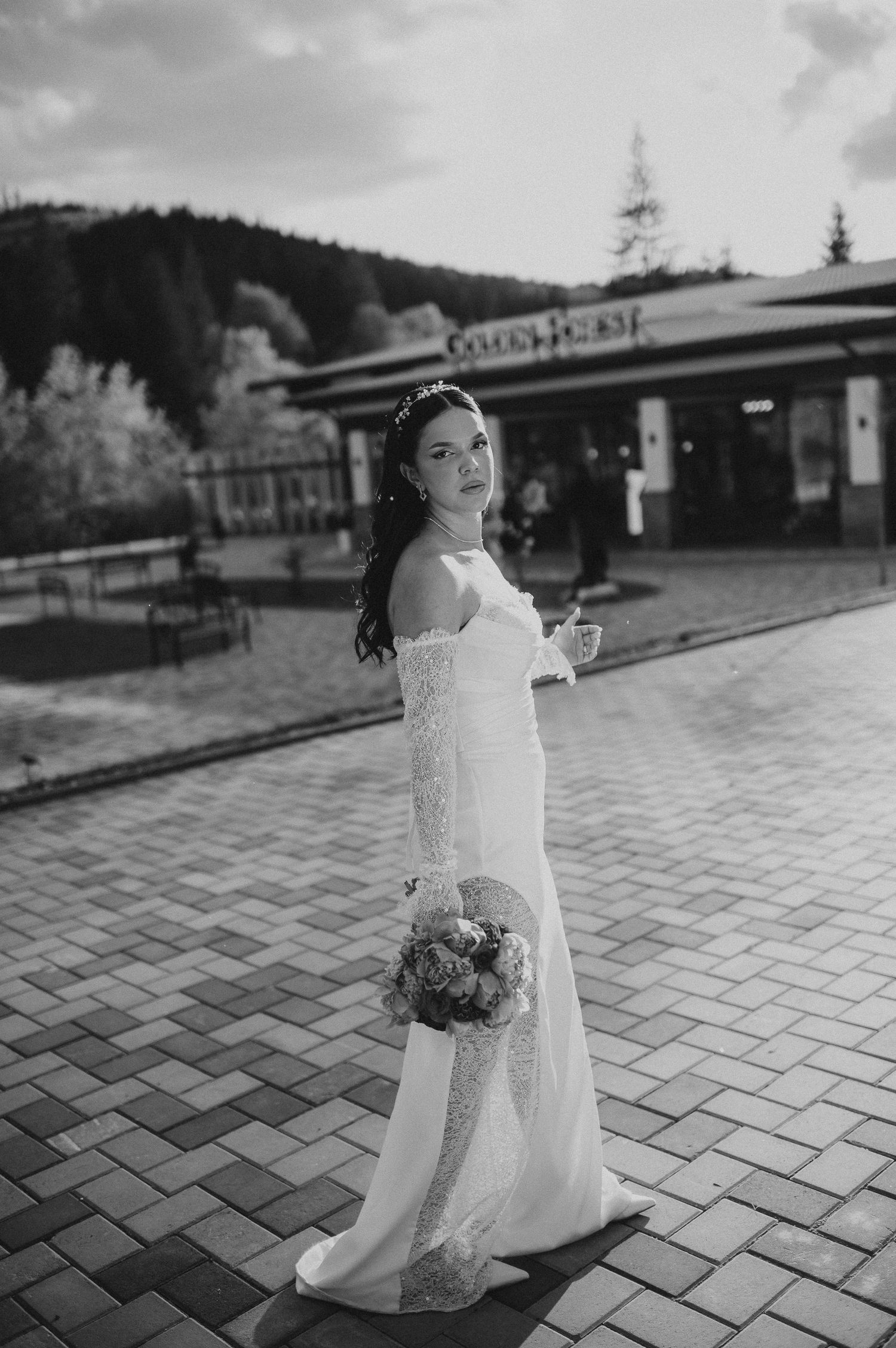 Black and white portrait of bride in off-shoulder wedding gown holding bouquet outside Golden Forest venue.