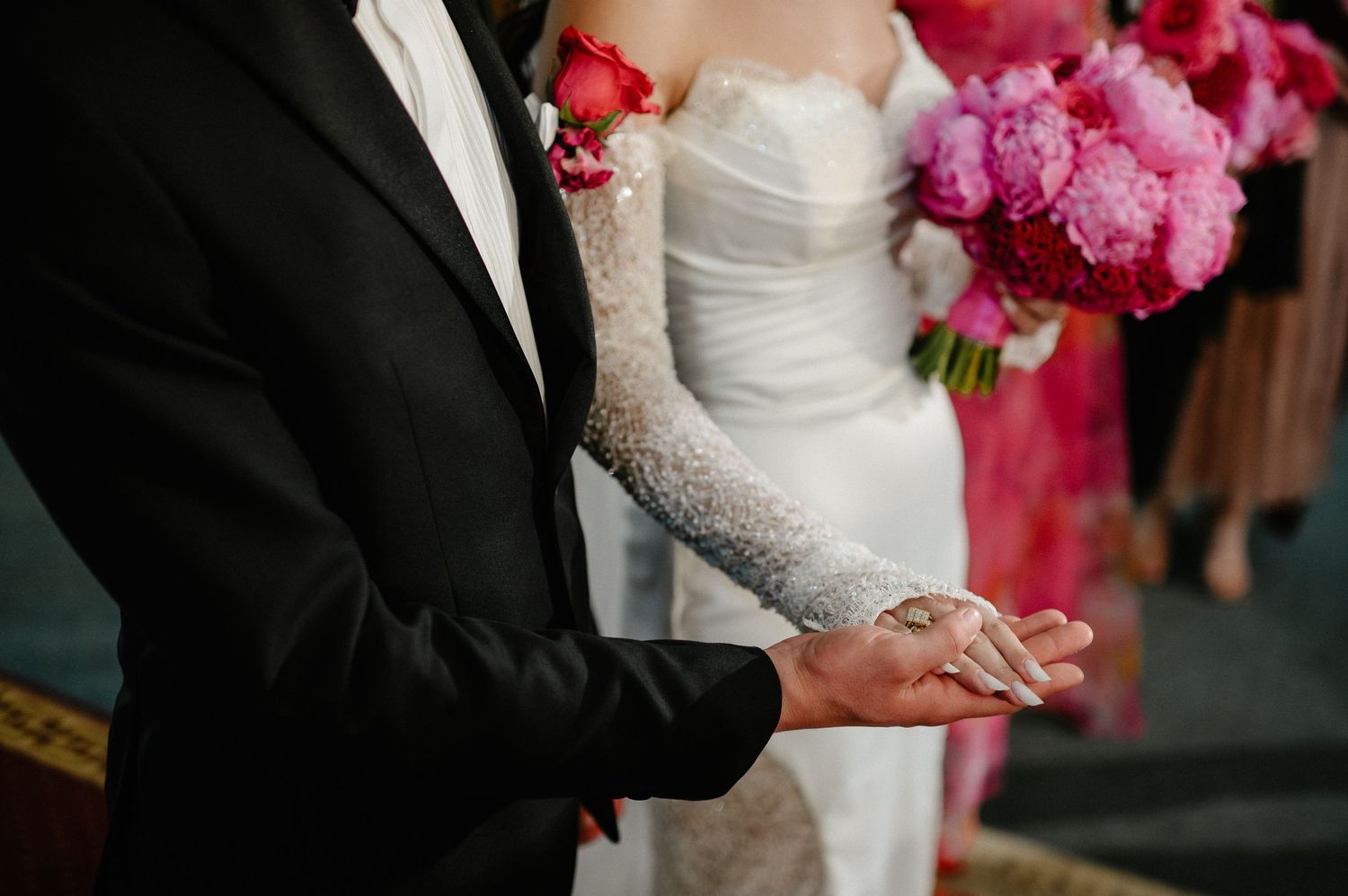 Groom in black tuxedo holds wedding rings while bride in lace gloves stands beside him.