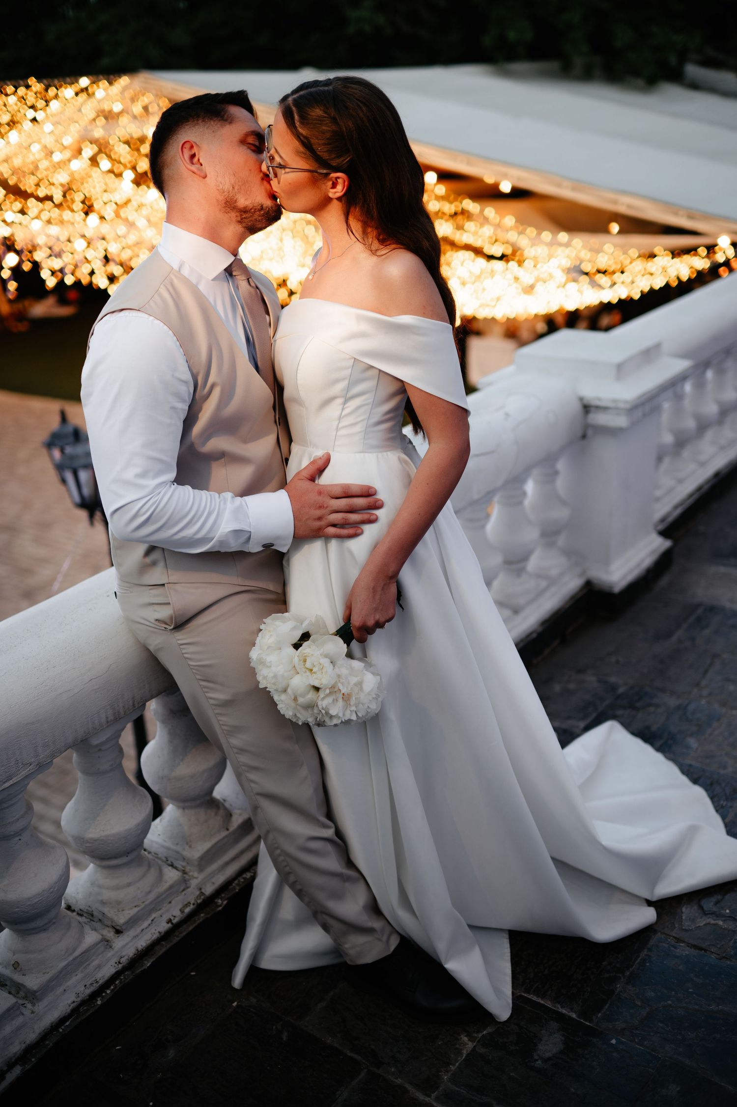 Bride and groom sharing a kiss on a balcony with glowing string lights at their evening wedding.