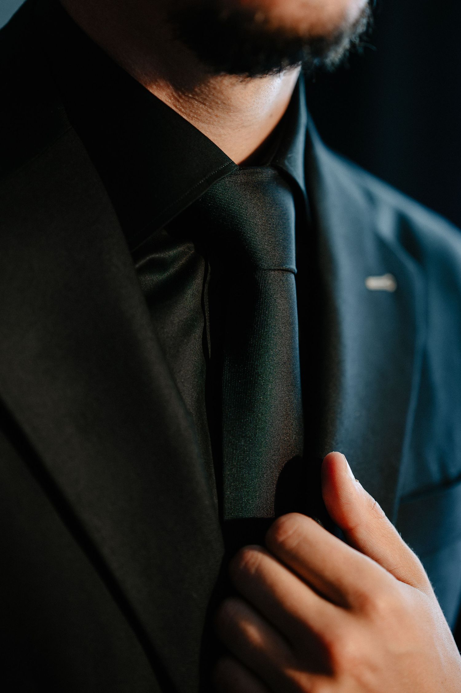 Close-up of a groom adjusting his black tie on a dark suit with lapel pin.