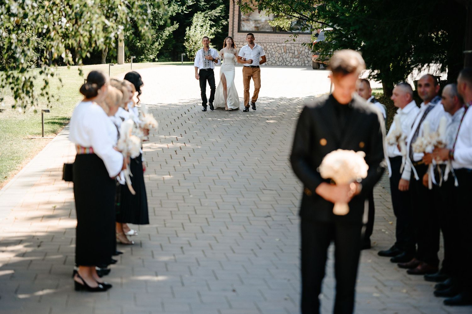 Wedding guests line brick pathway as groom holds white bouquet during ceremony.