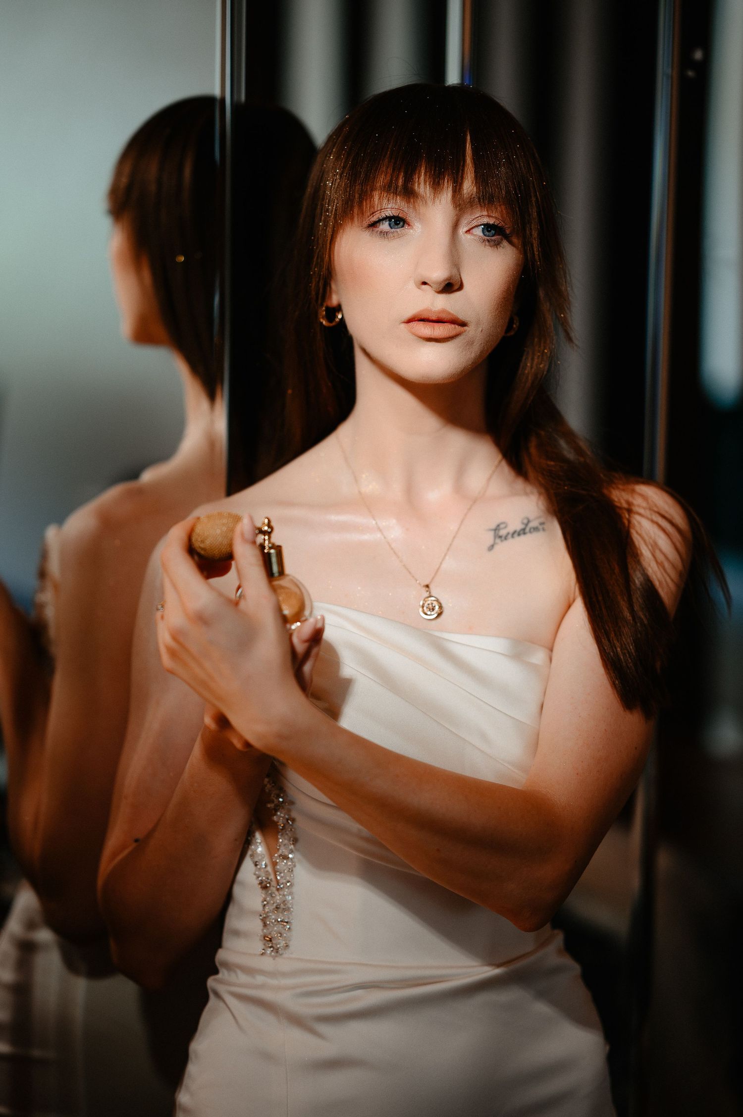 Bride in strapless white dress applies perfume while reflected in mirror with gold necklace.
