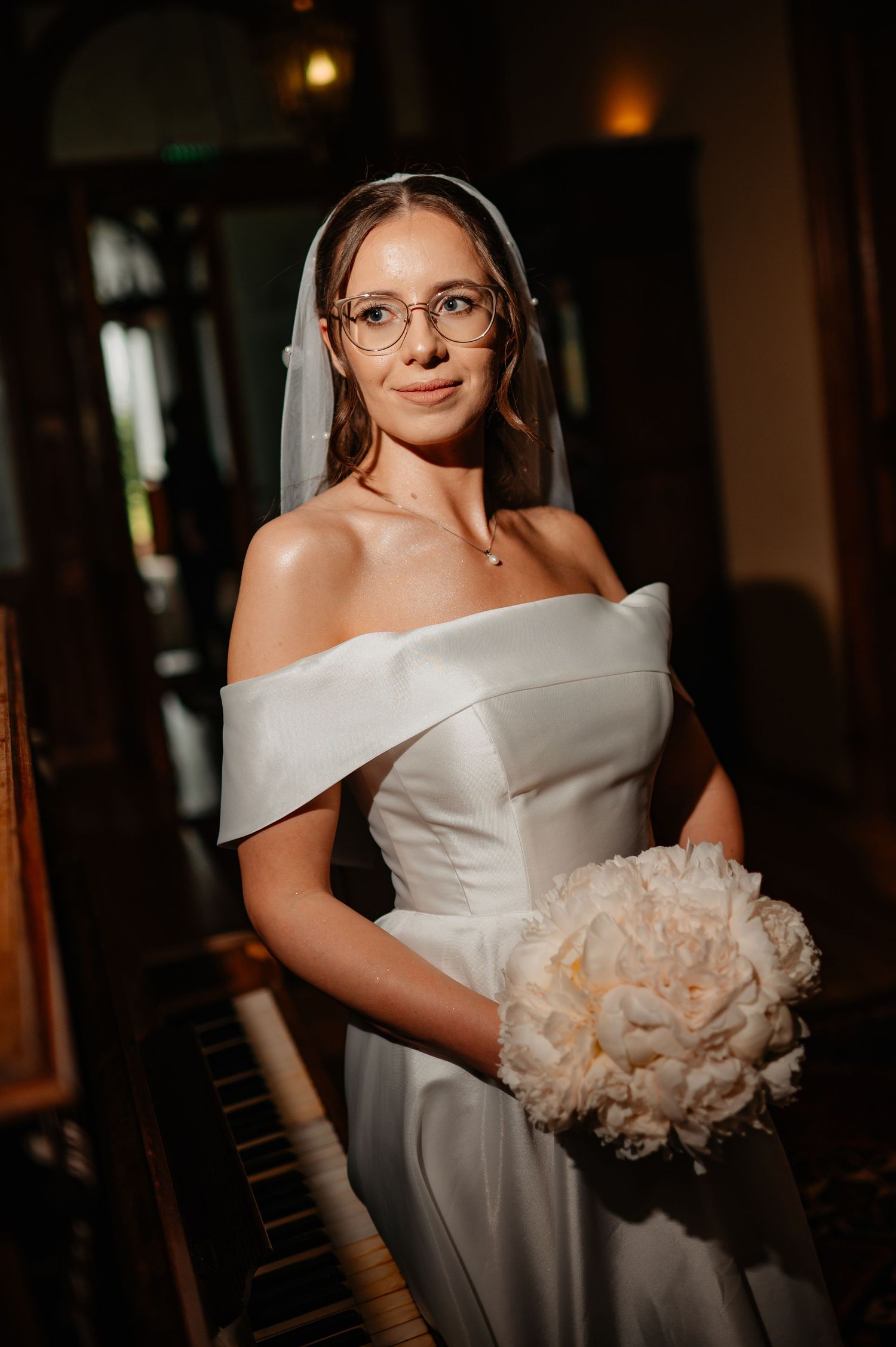 Bride wearing glasses and off-shoulder white gown holds white peony bouquet in dark interior.