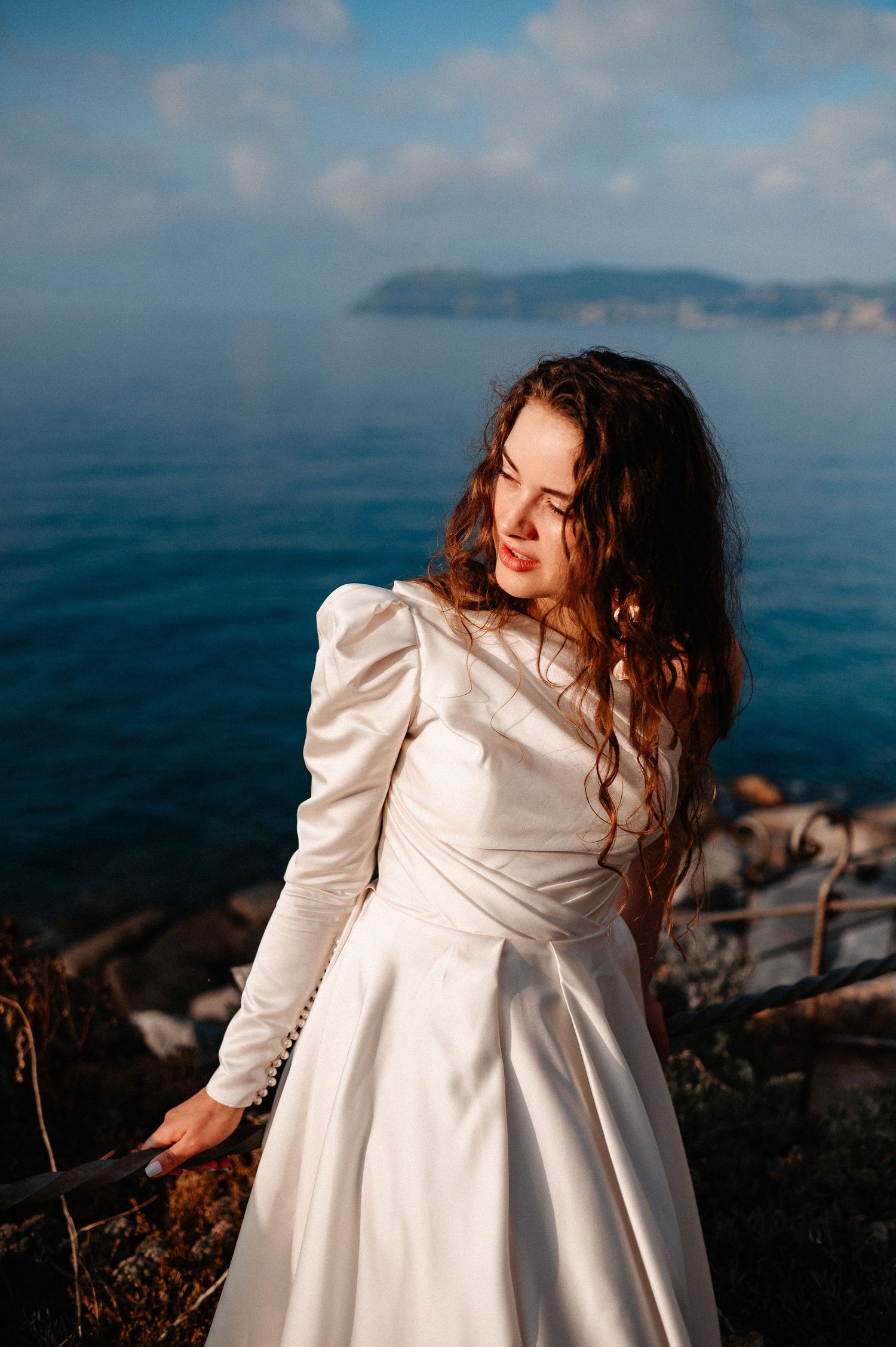 Curly-haired bride in white satin gown smiling softly by seaside with island in background.