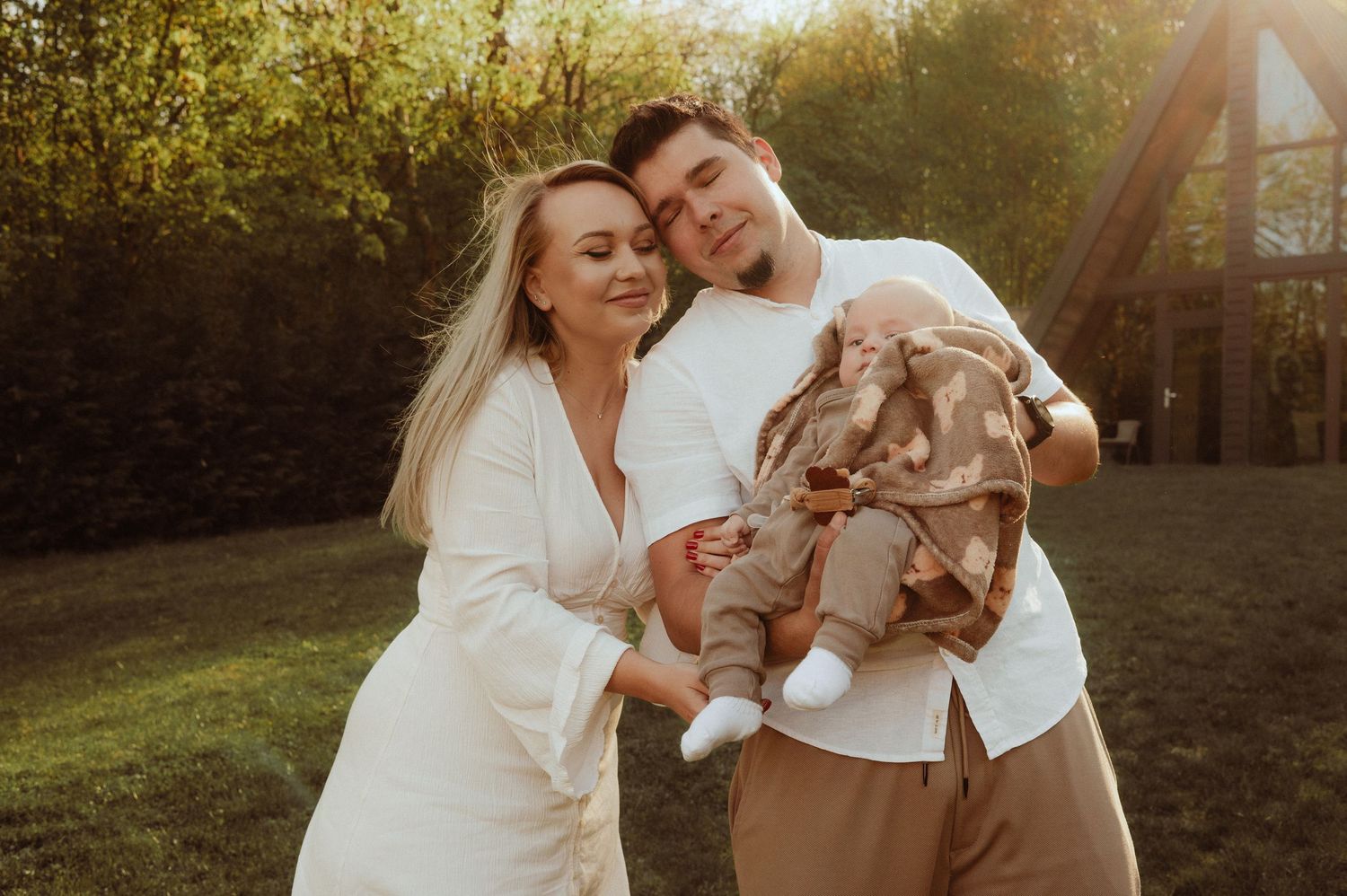 Happy couple smiling and cuddling their newborn baby outdoors in warm golden afternoon sunlight.