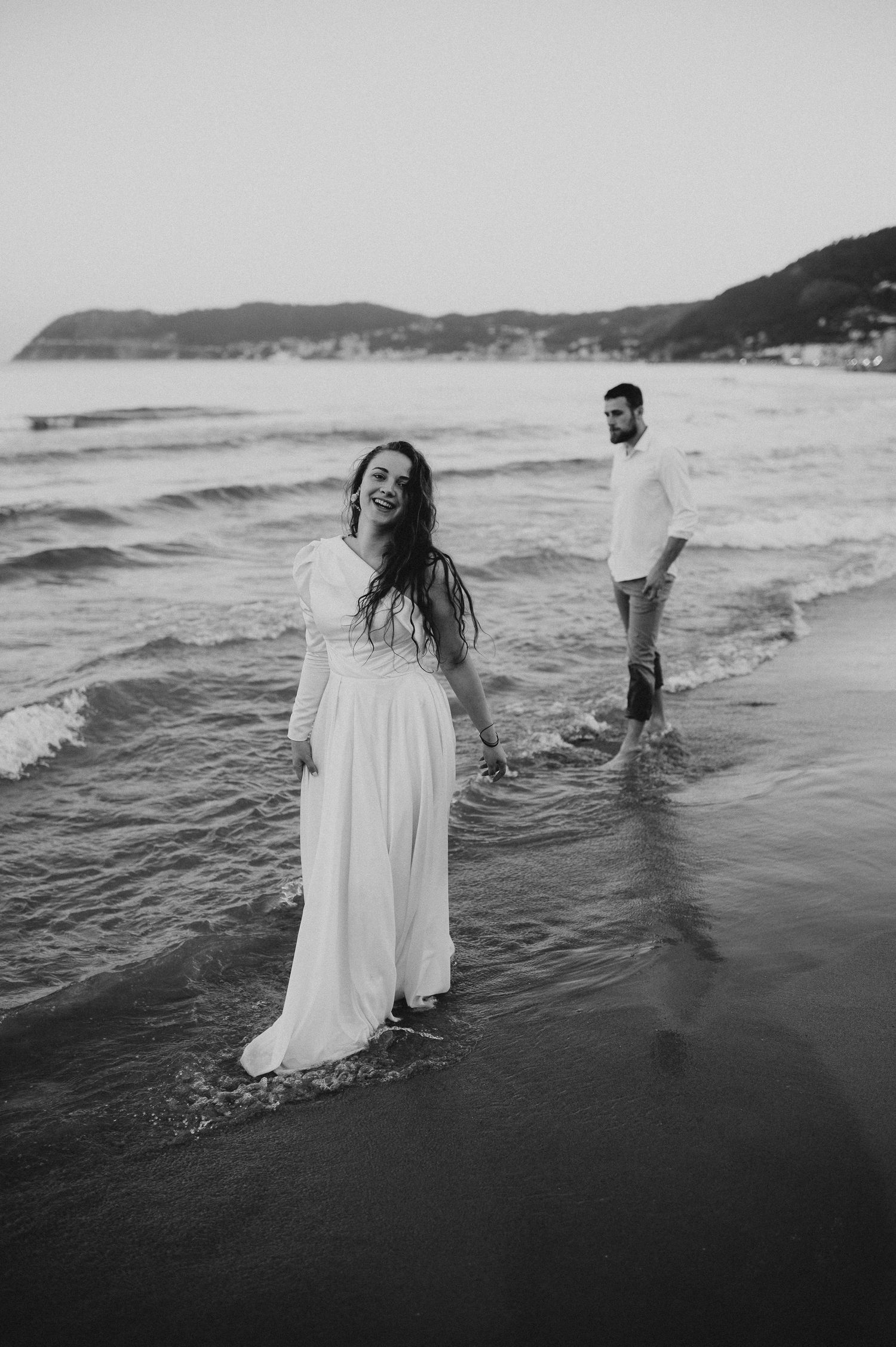 Black and white photo of smiling bride in white dress on beach with groom.