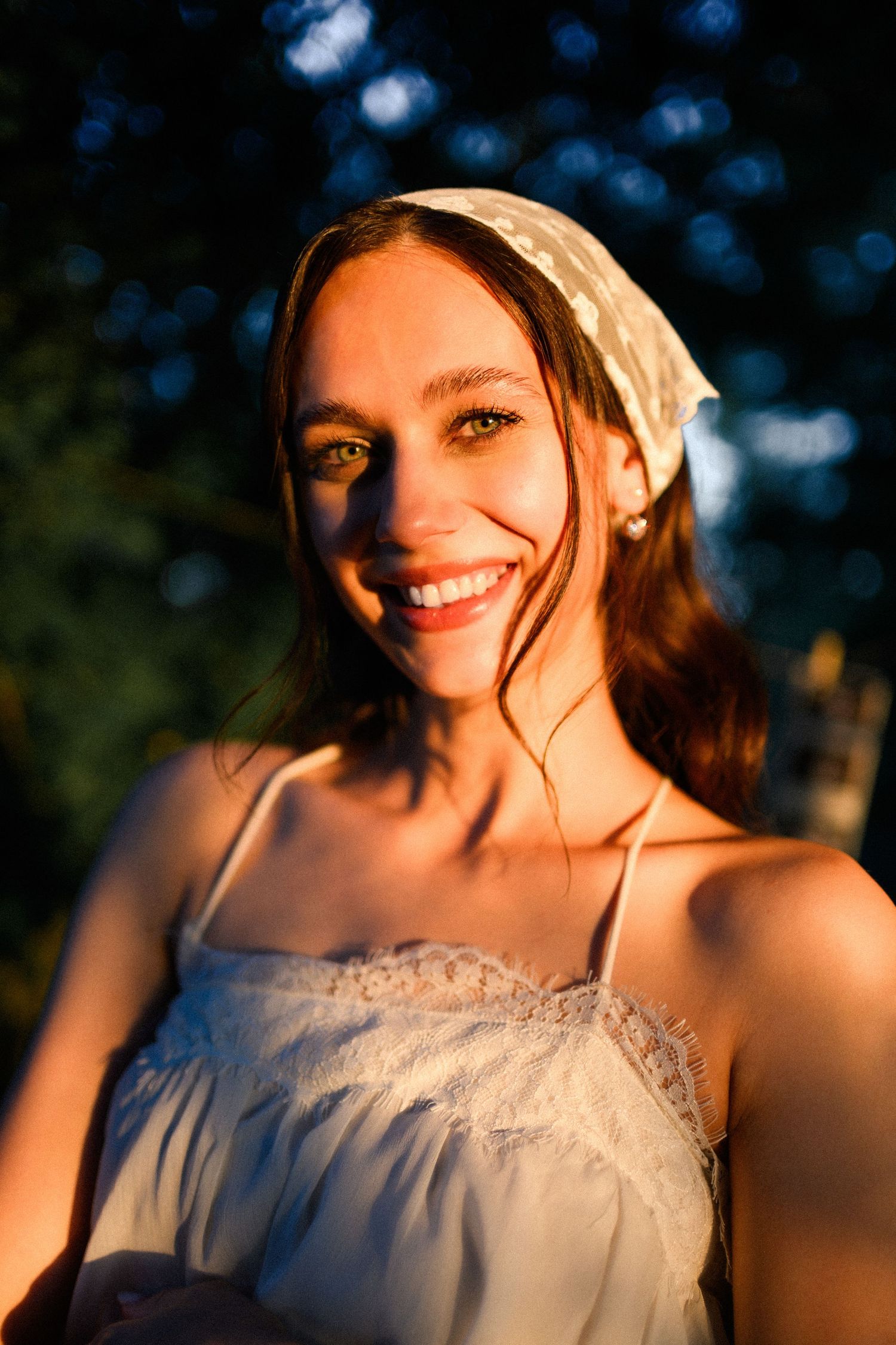 Smiling young woman in white sundress and headscarf bathed in warm golden sunset light outdoors.