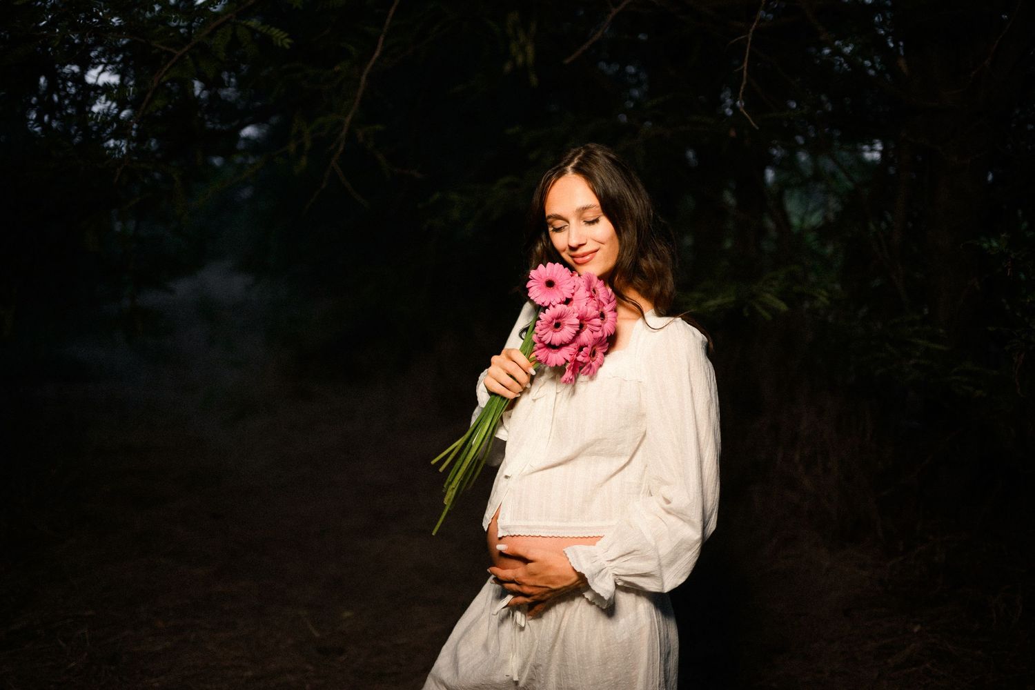 Pregnant woman in white dress holding pink flowers, smiling outdoors in dark forest setting.