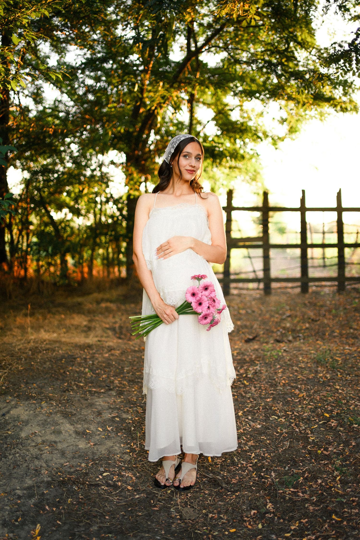 Pregnant woman in white dress holding pink flowers outdoors near wooden fence with lush green trees in background.