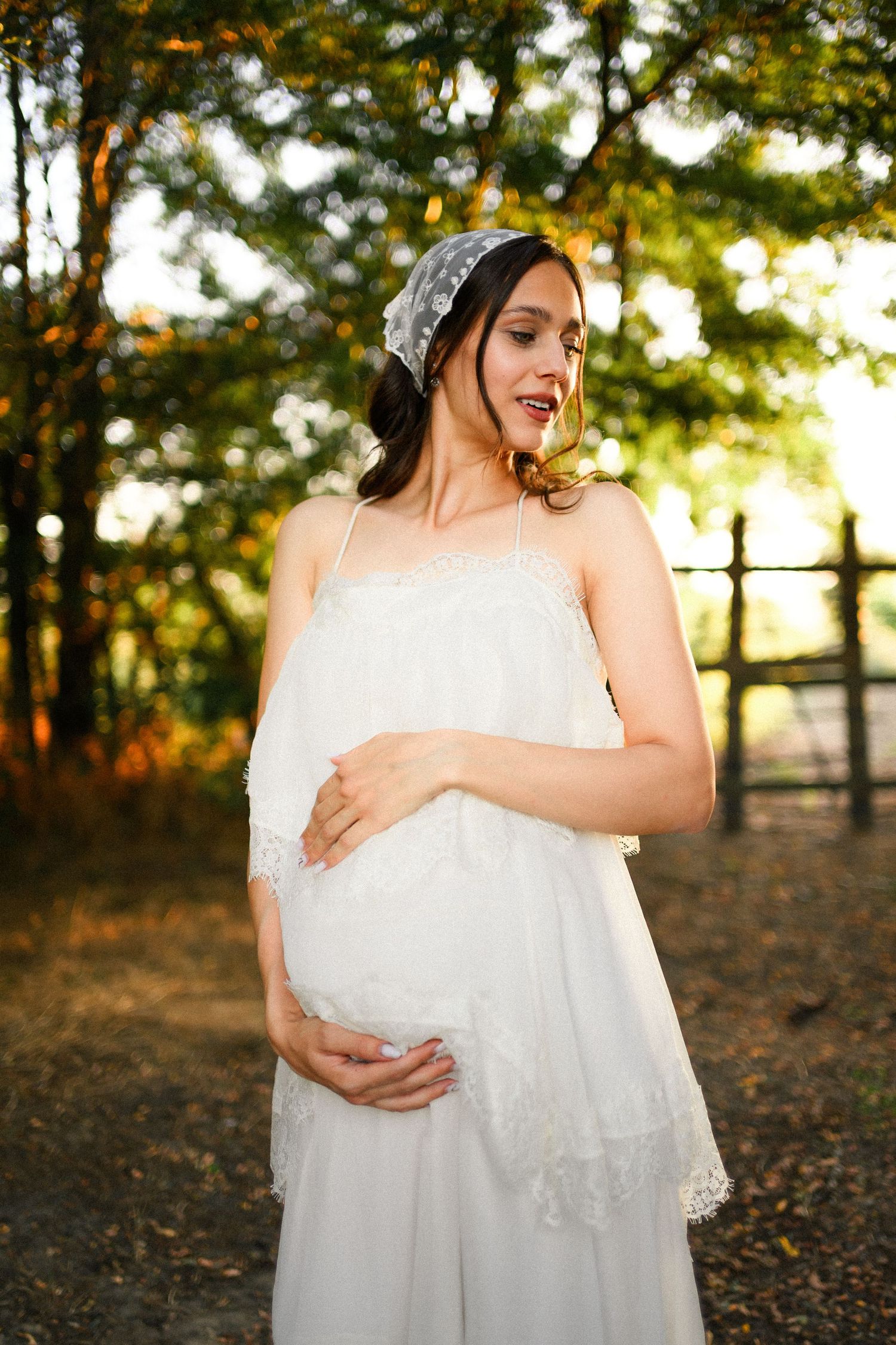 Pregnant woman in white lace dress and headpiece cradling her baby bump outdoors in golden sunlight.