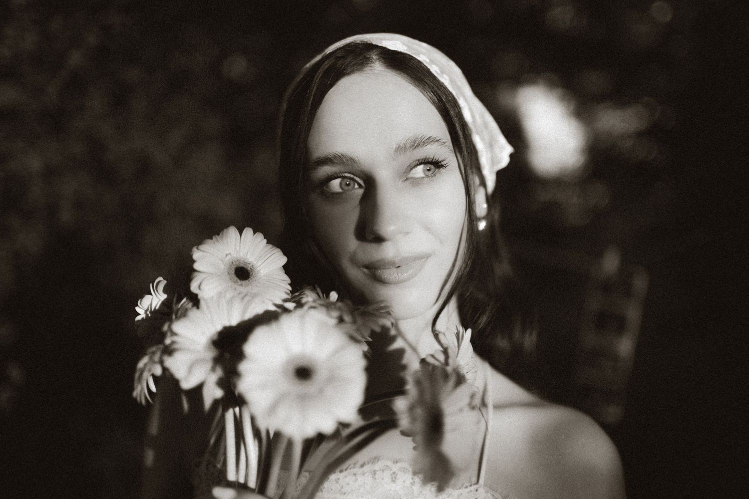 Black and white portrait of a bride holding white gerbera daisies, wearing a veil, with a soft bokeh background.