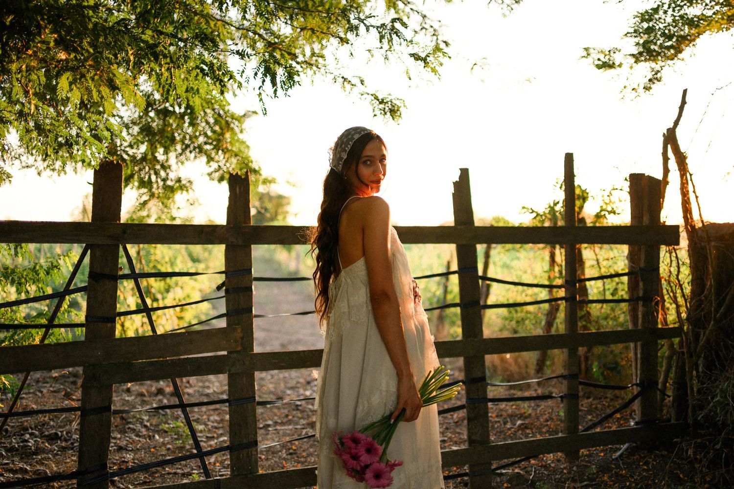Bride in white dress holding pink flowers stands by rustic wooden fence at golden hour sunset.