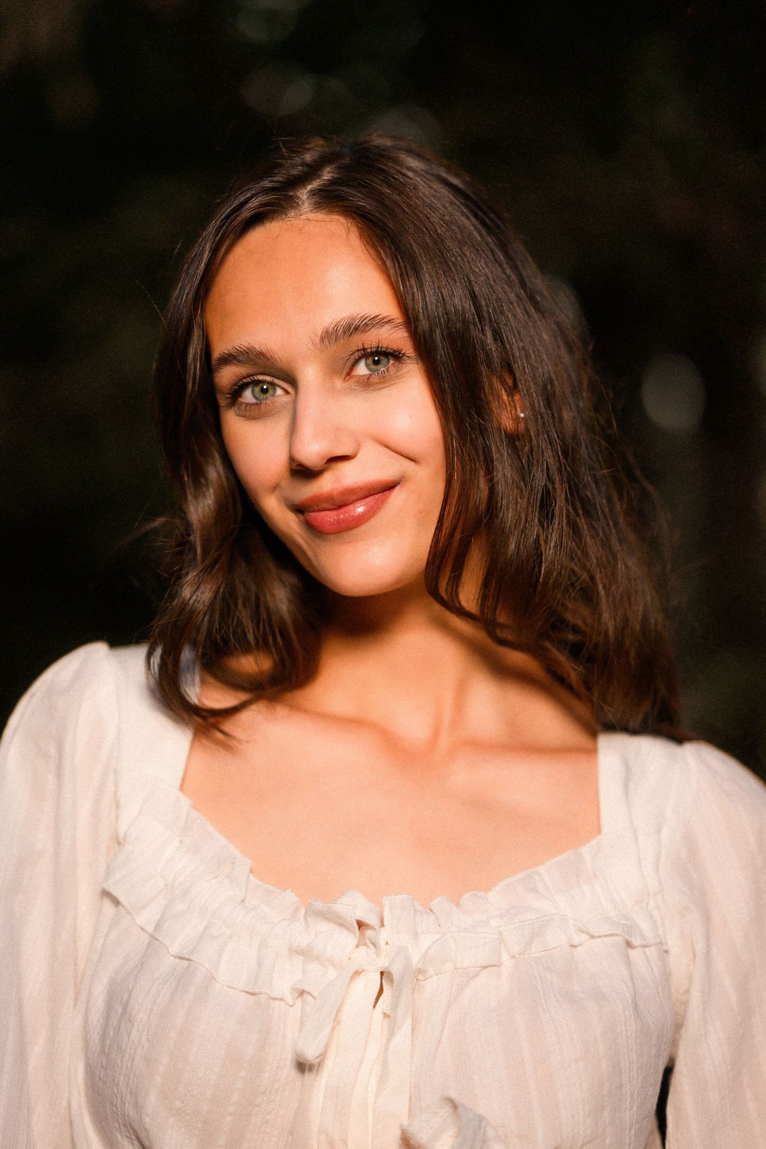Young woman with brown wavy hair smiling softly, wearing a white ruffled blouse outdoors in warm golden light.